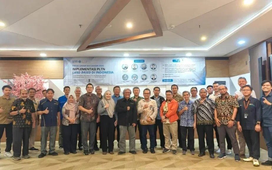 Group of around 20 people standing together indoors, posing for a photo at a conference or seminar. There is a large banner behind them with text and logos, and a pink tree on the left side of the scene.