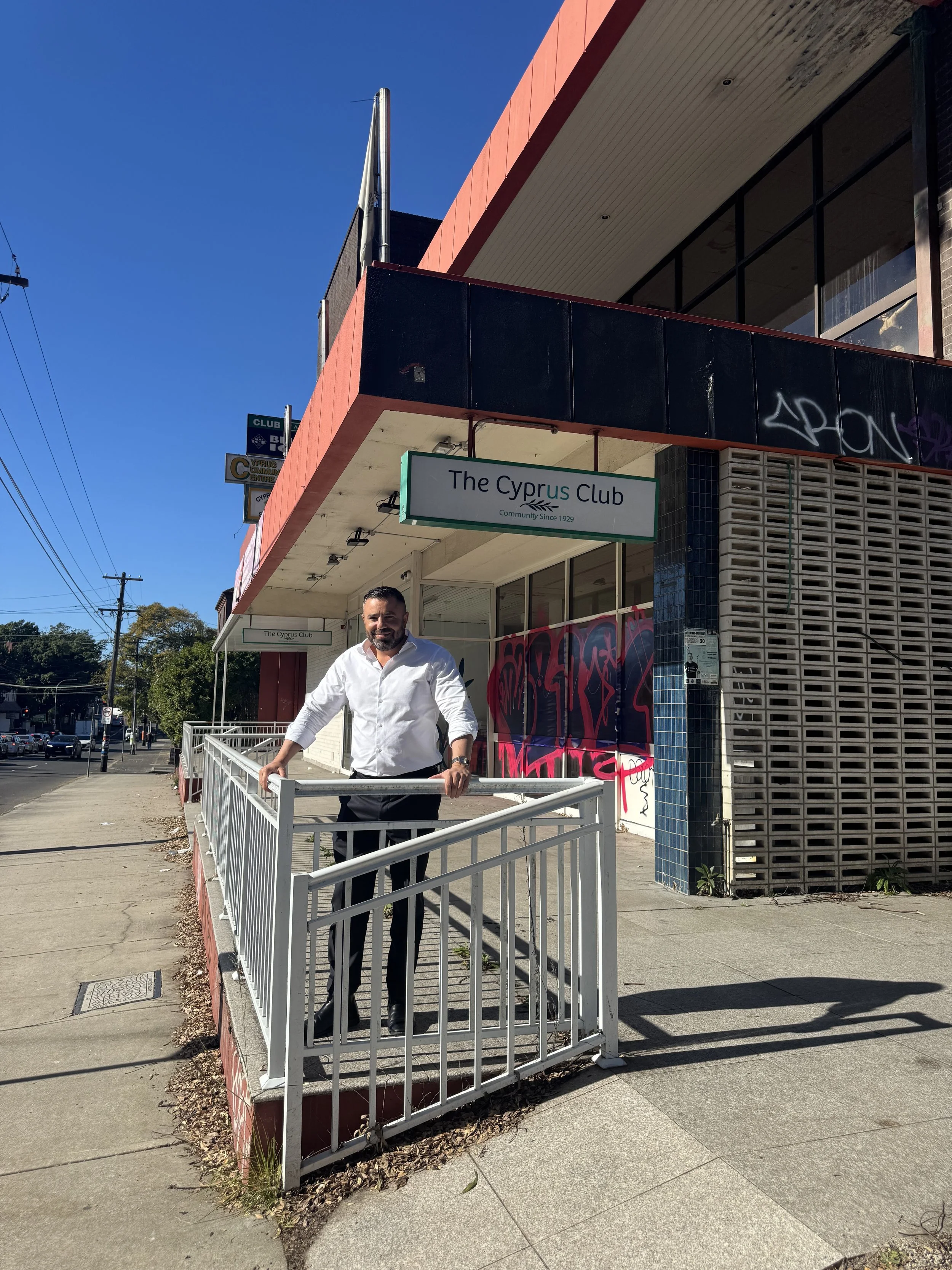 A man standing on a sidewalk in front of a building with a sign that reads 'The Cyprus Club,' with blue sky and power lines in the background.