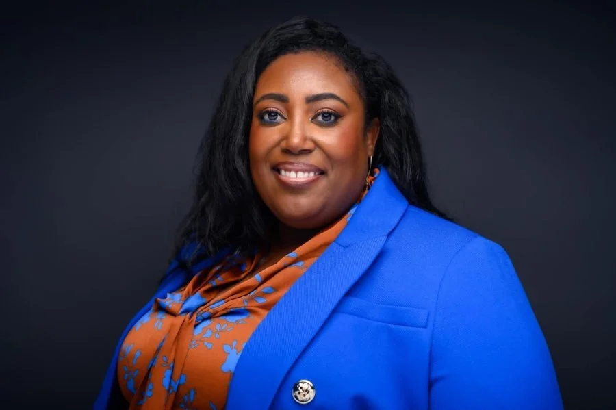 A woman with dark hair, wearing a blue blazer and an orange patterned scarf, smiling against a dark background.