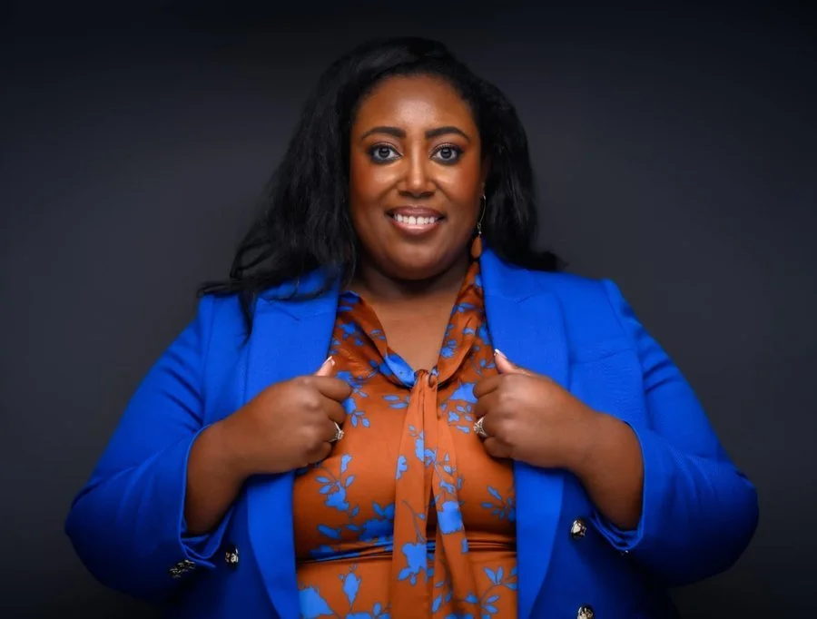 A smiling woman with dark hair, wearing a bright blue blazer over a brown blouse with blue floral patterns, adjusted her blazer with both hands, standing against a dark background.