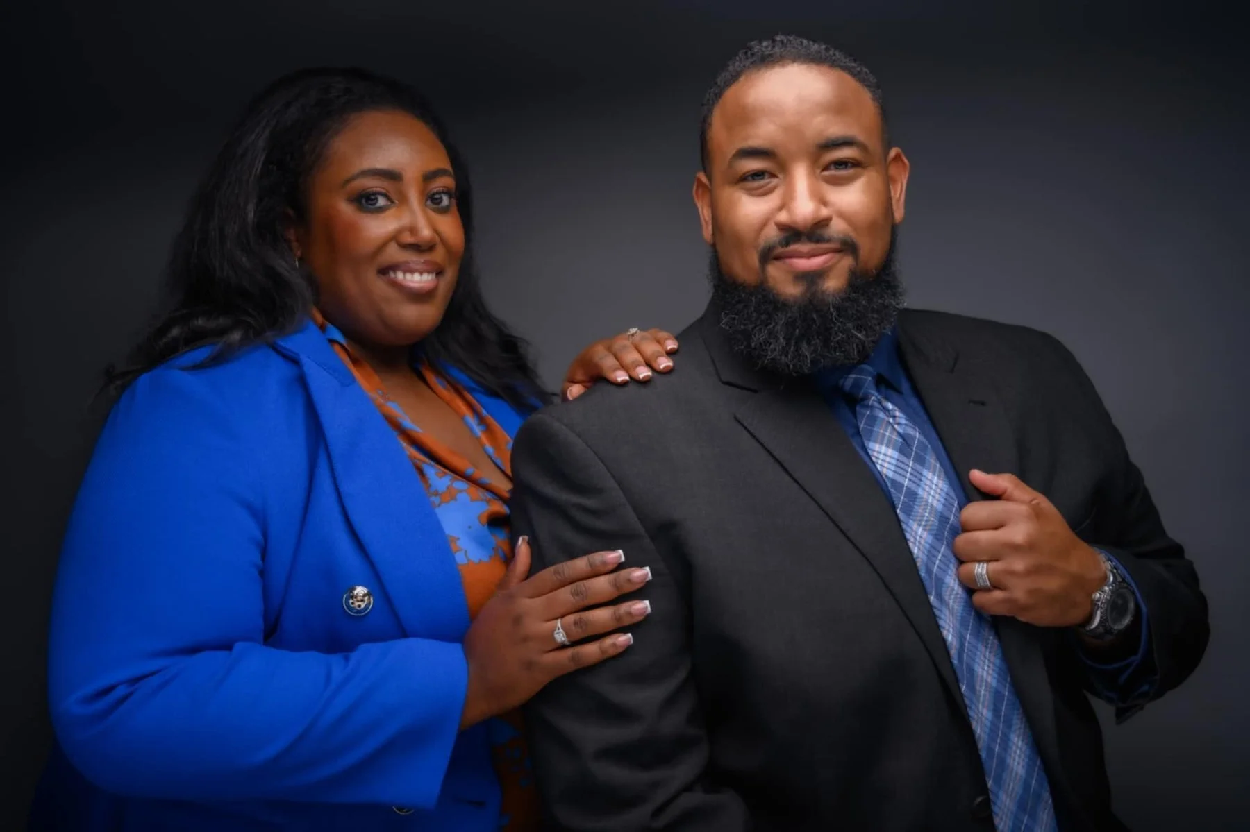 A smiling Black woman with black hair, wearing a bright blue blazer and a patterned orange and blue top, standing beside a smiling Black man with a beard, wearing a dark suit, blue shirt, and plaid tie. They pose together against a dark background, with the woman resting her hand on the man's shoulder.
