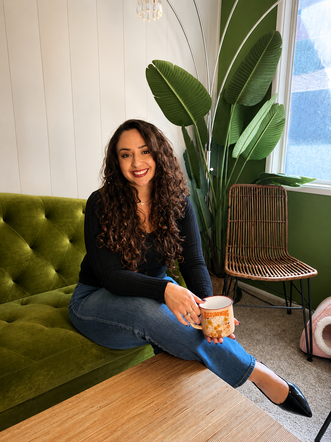 A woman with curly brown hair sitting on a green velvet couch, holding a mug, smiling indoors with a large green plant, wicker chair, and window in the background.