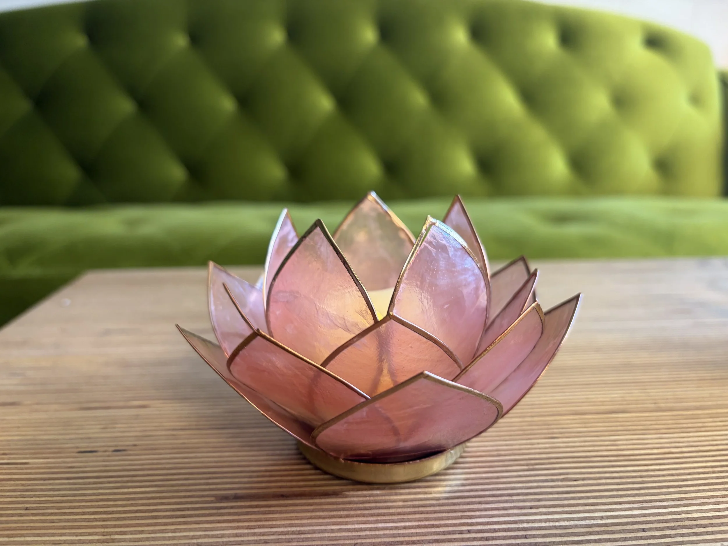 Pink lotus-shaped candle holder with a lit candle inside, placed on a wooden surface with a green tufted background.