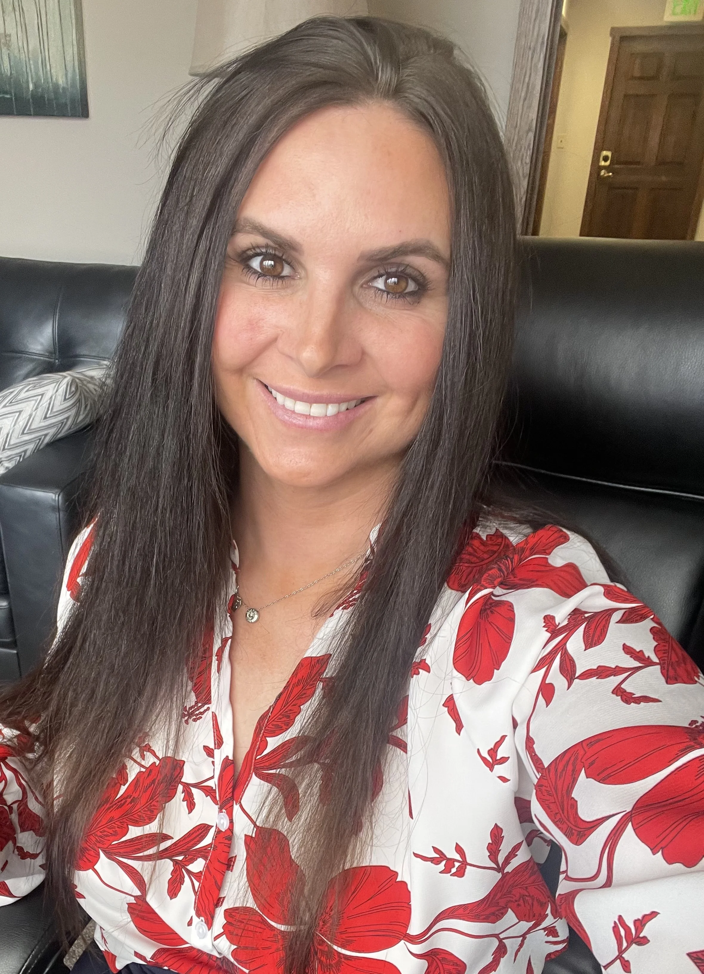 Close-up of a woman with long dark hair, light skin, brown eyes, smiling, wearing a white blouse with red floral patterns, sitting on a black leather chair with a wooden door and a painting in the background.