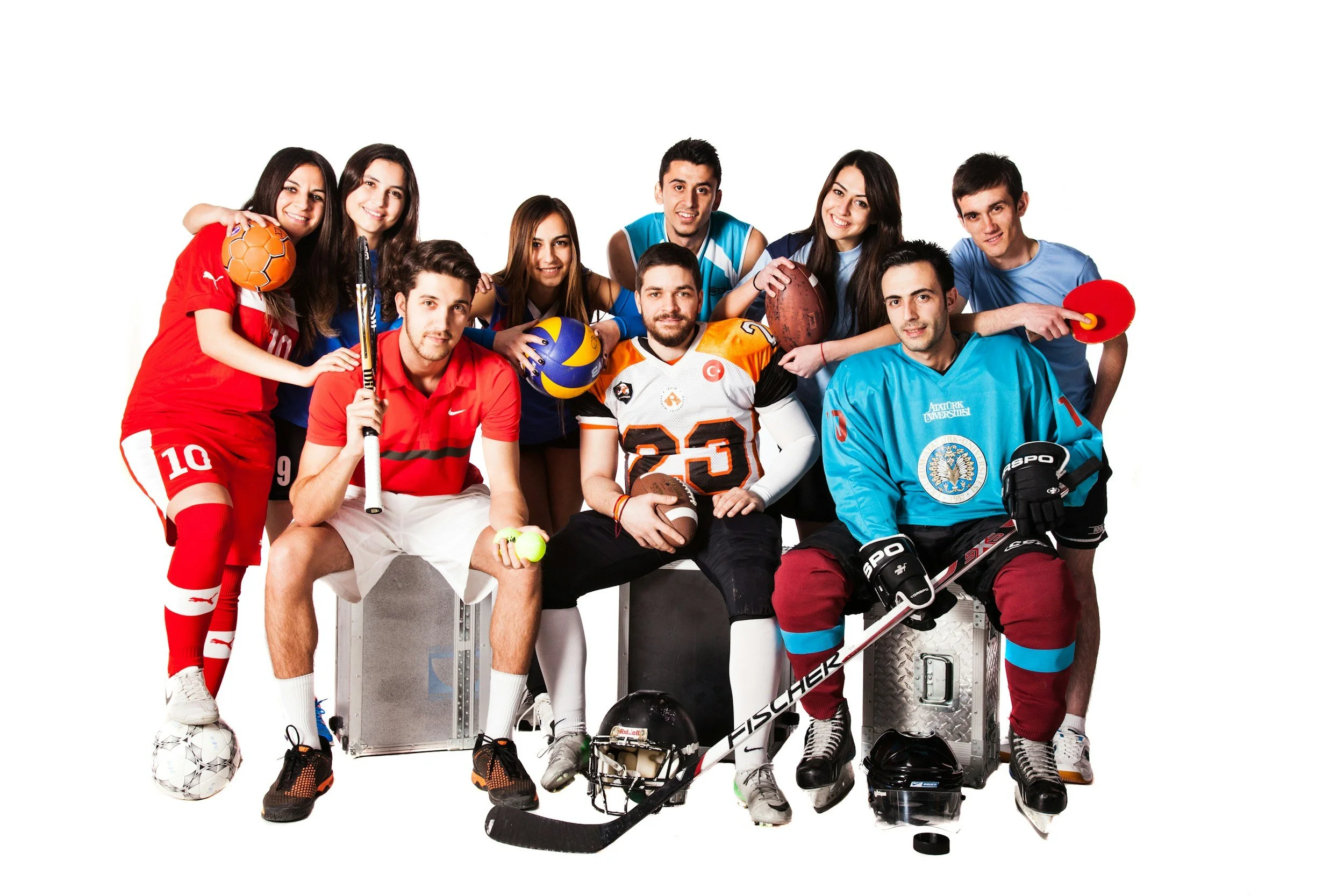 Group of young athletes, including basketball, volleyball, football, and hockey players, posing together with sports equipment against a white background.