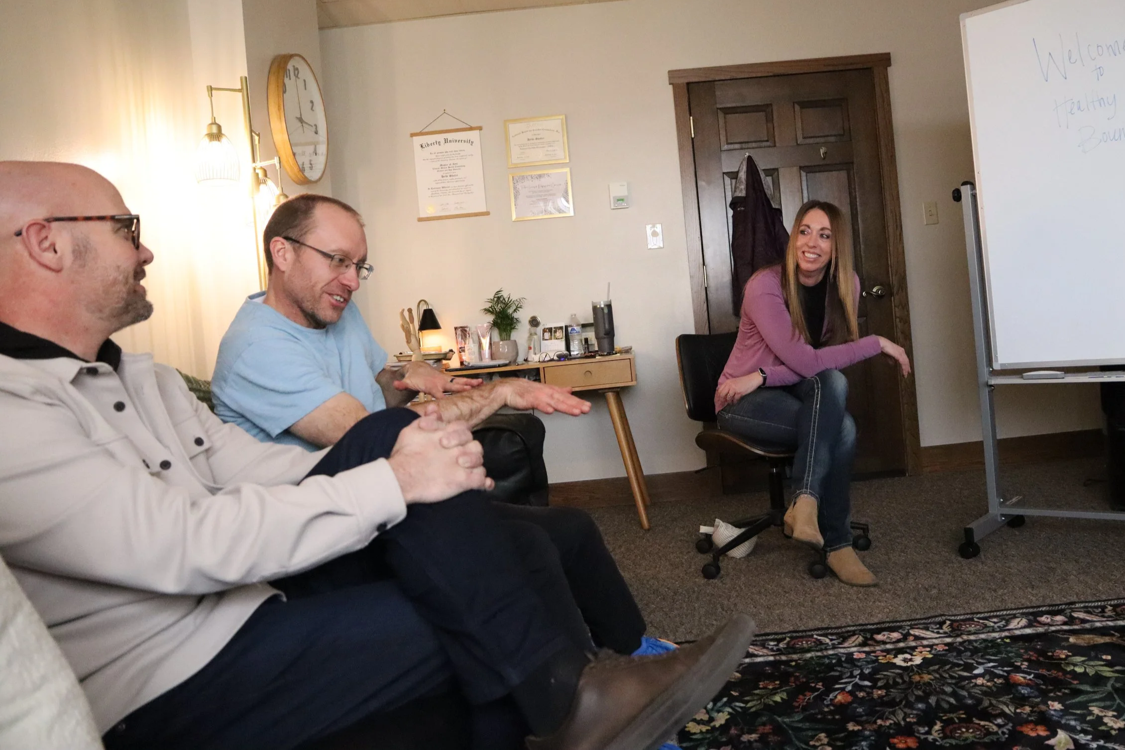 Four adults sitting in a room, engaged in a discussion or meeting. A woman is sitting in a chair near a whiteboard, smiling, while two men are seated on the left side of the image, and one of them is gesturing with his hands. The room has a wooden door, framed documents on the wall, a small desk with items, and a patterned rug.