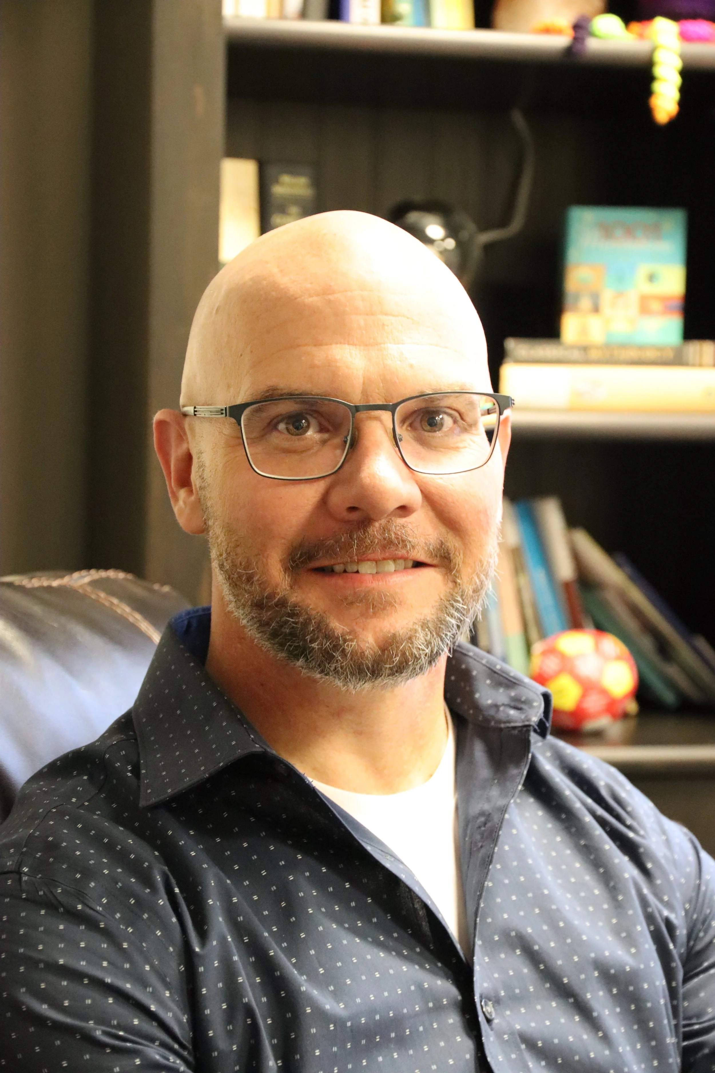A man with a bald head, glasses, and a beard smiling, sitting indoors with bookshelves in the background. The man is wearing a dark, patterned button-up shirt.