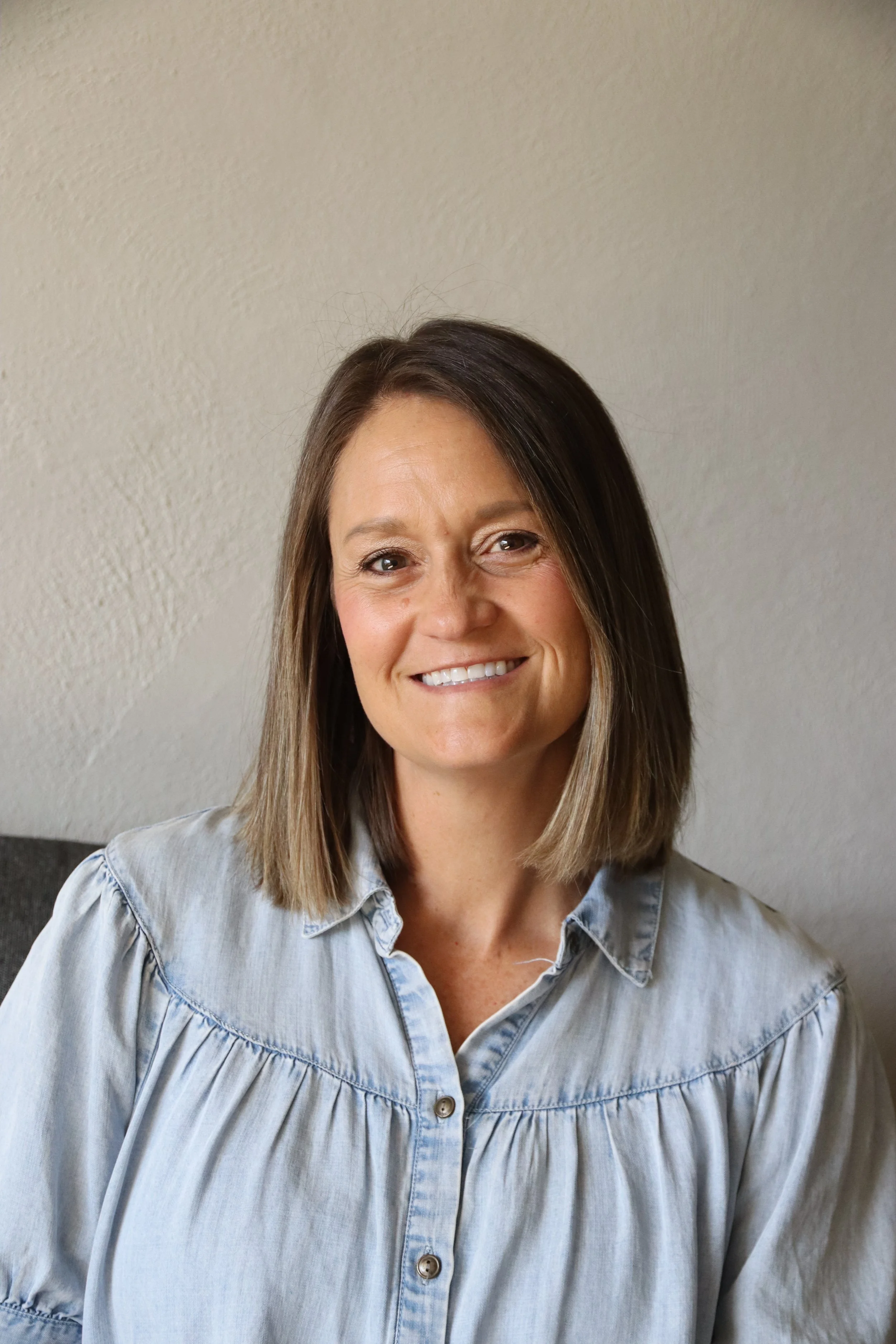 A woman with medium-length brown hair and a light blue denim shirt smiling, standing against a plain wall.