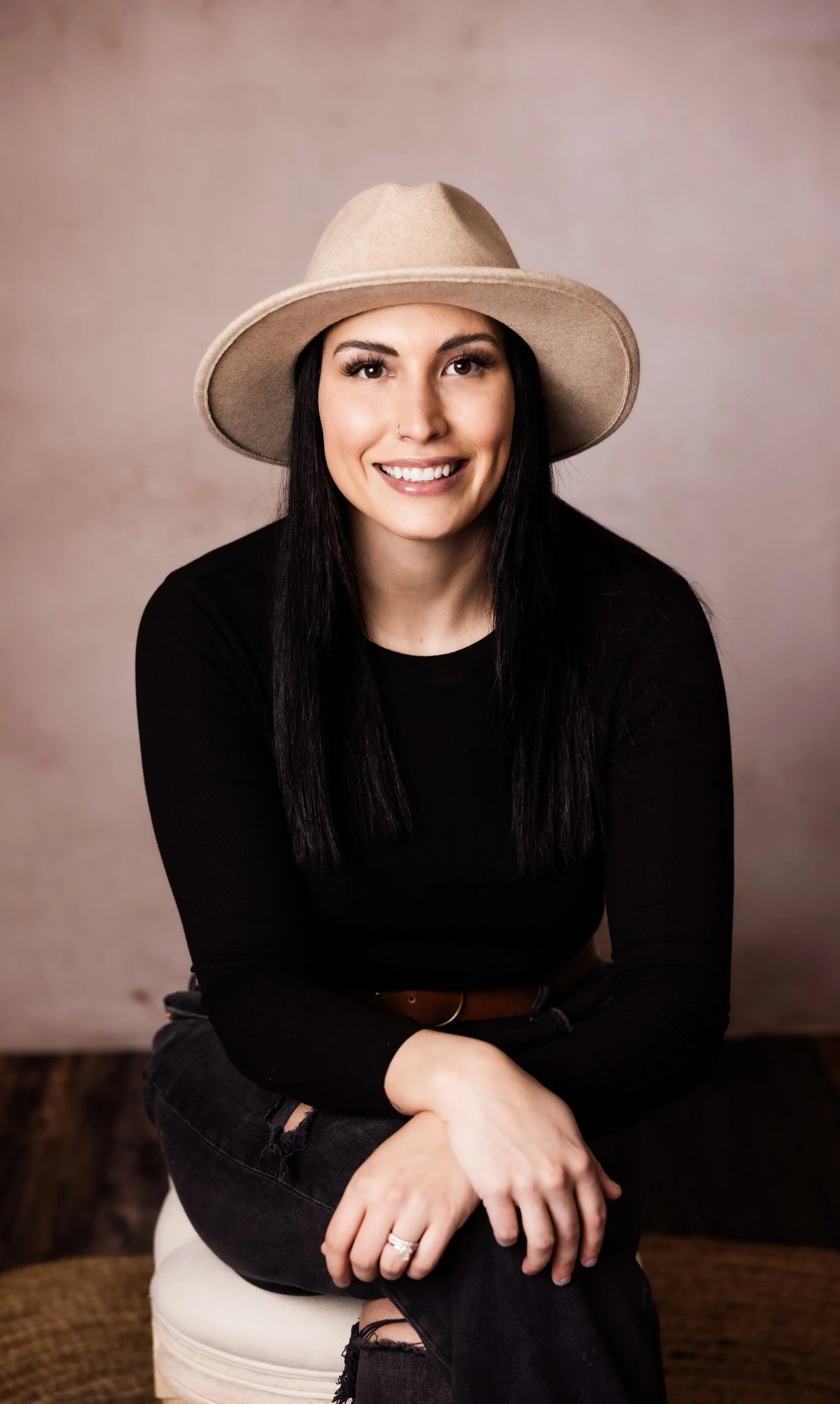 A woman with long black hair and a septum piercing, smiling and wearing a wide-brimmed beige hat and a black long-sleeve shirt, sitting on a beige chair against a neutral background.