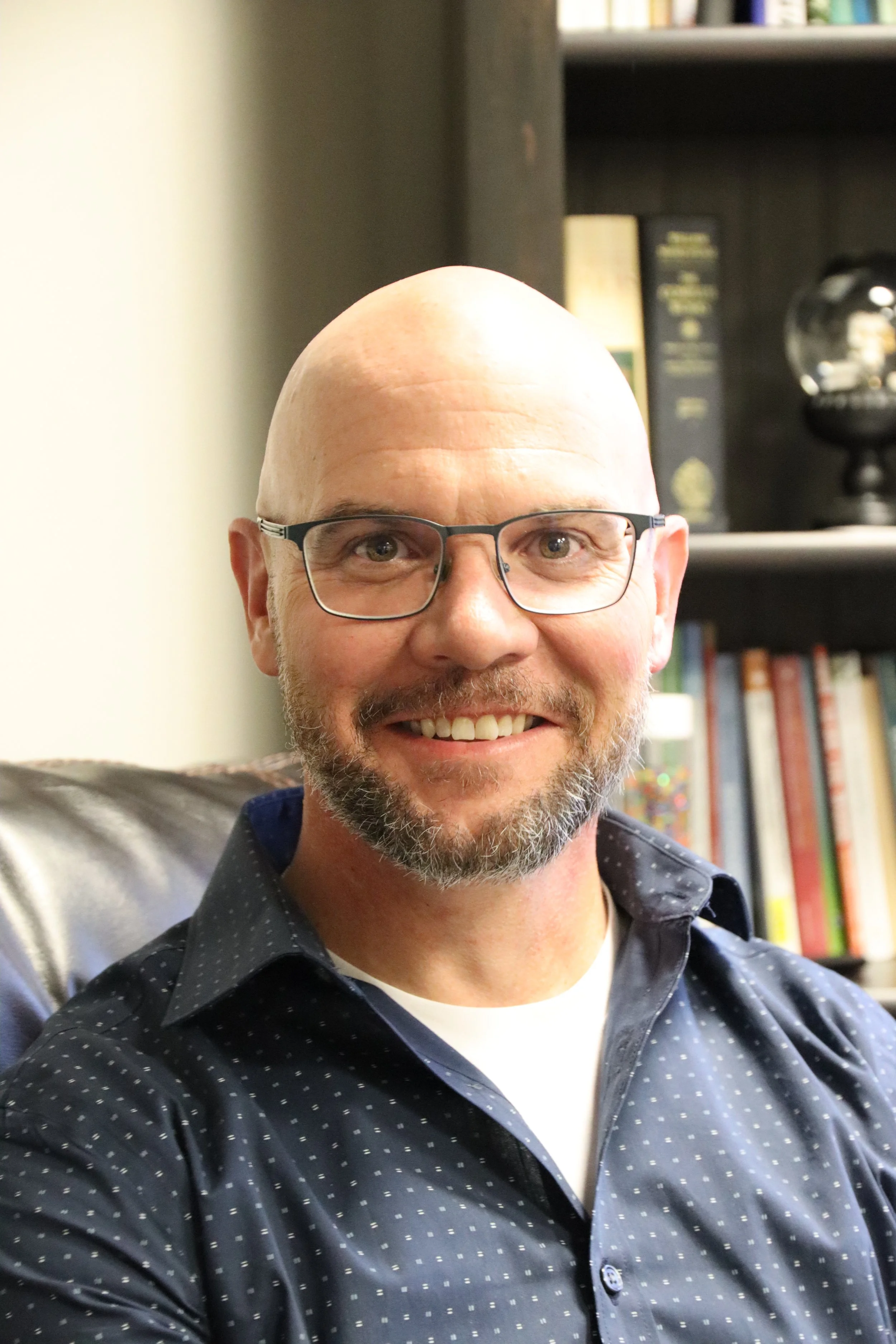 A smiling bald man with glasses taking a selfie inside a room with a bookshelf in the background.