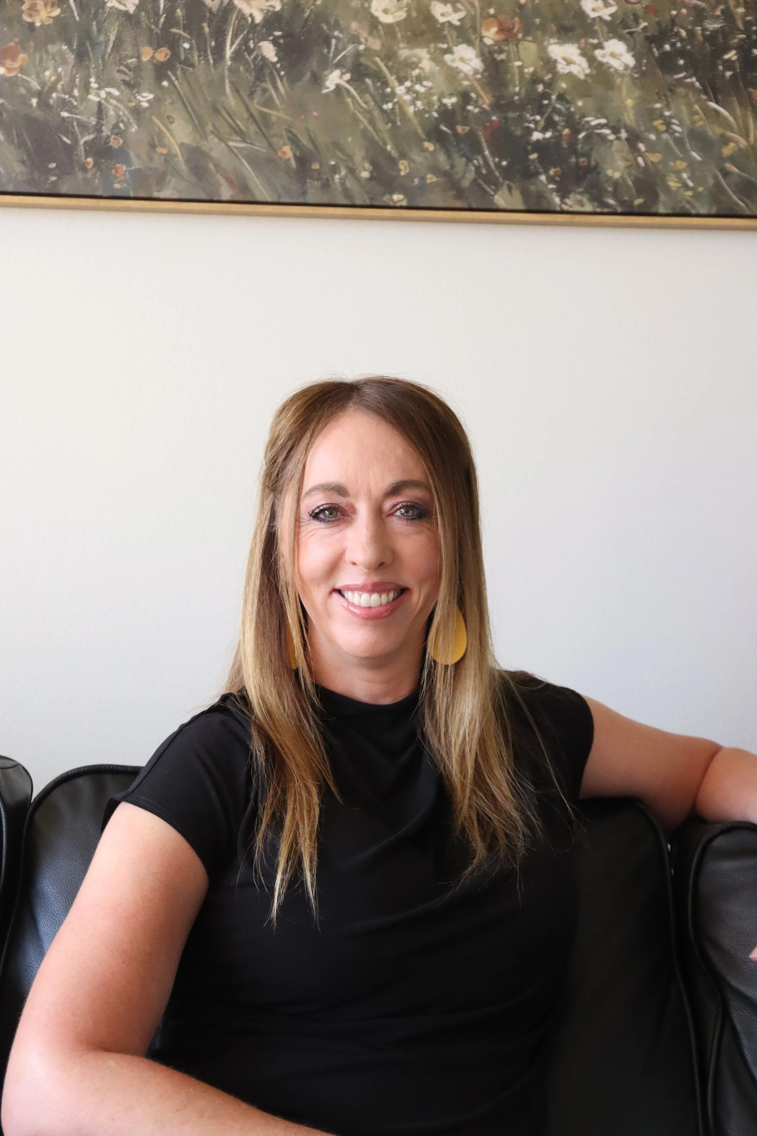 A woman with long light brown hair, wearing a black shirt and yellow earrings, smiling while sitting on a black leather couch in a room with a plain white wall and a framed abstract painting hanging above her.