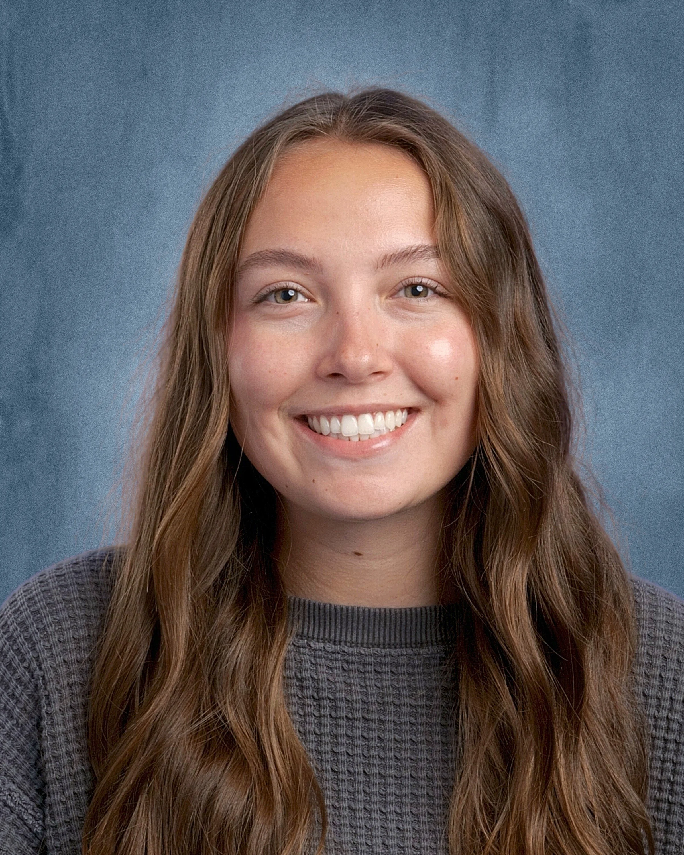 Close-up portrait of a young woman with long, wavy brown hair, smiling, wearing a gray knit sweater, against a blue background.