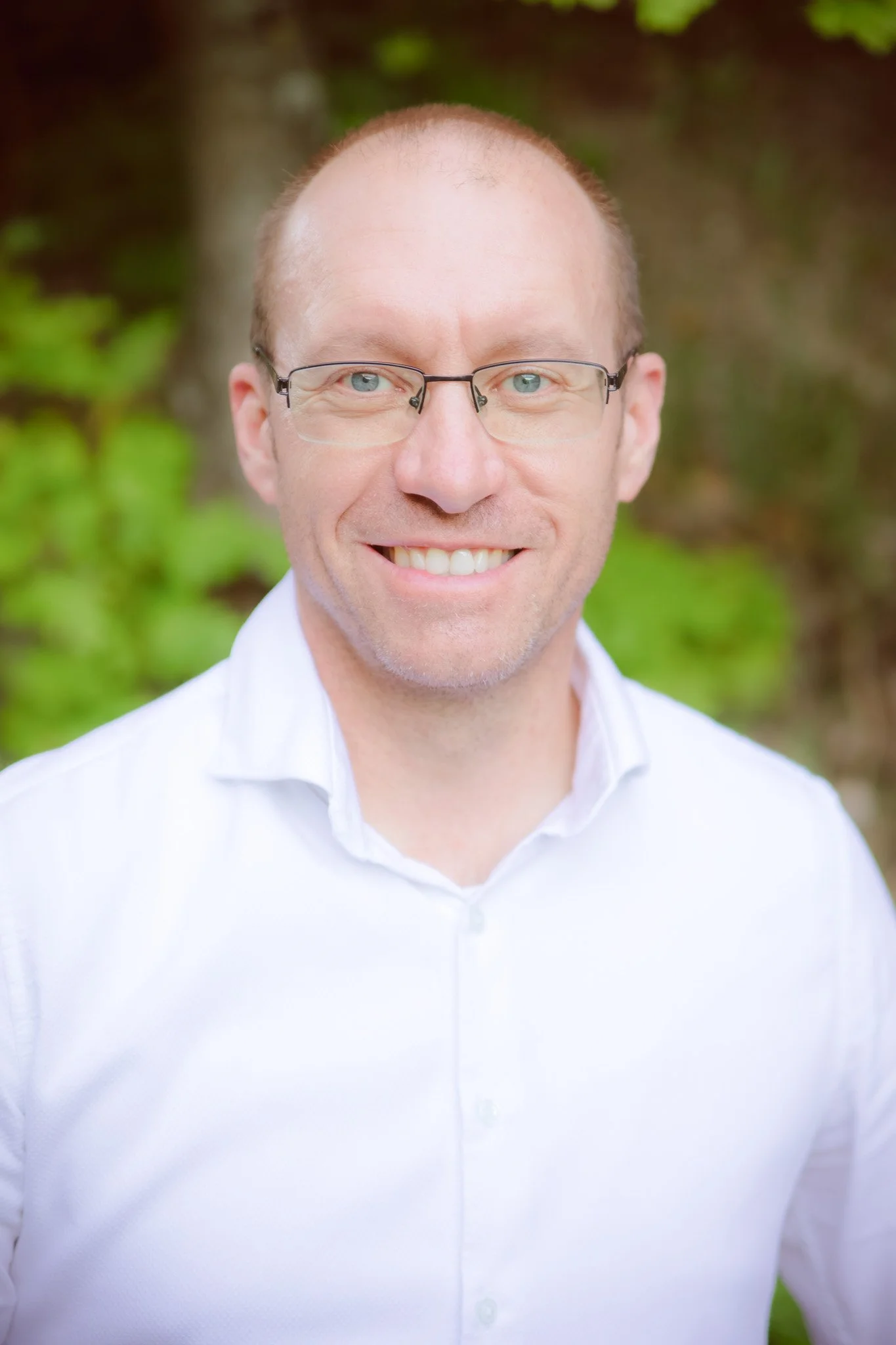 Close-up of a smiling man with short hair, wearing glasses and a white shirt, outdoors with blurred green foliage in the background.