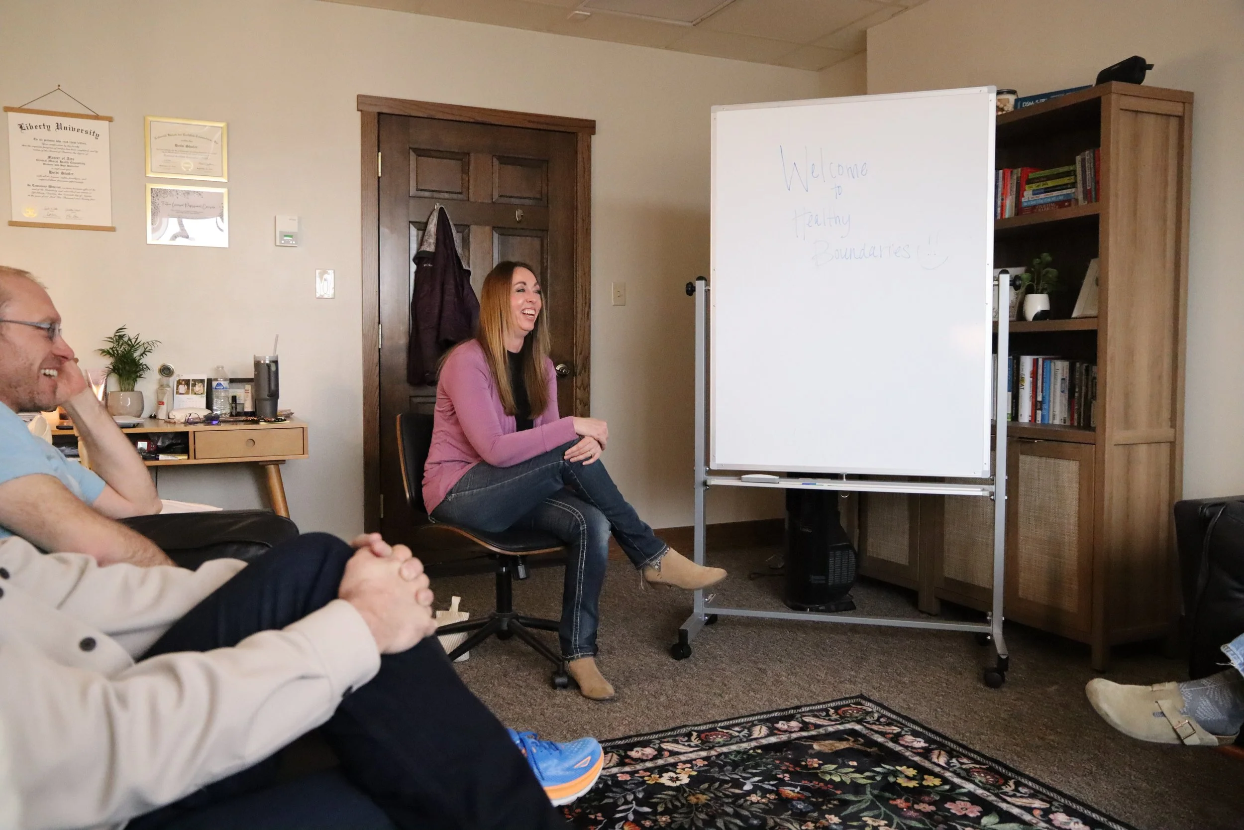 People sitting in a room, laughing and smiling during a discussion or presentation, with a woman seated near a whiteboard that reads 'Welcome to healthy boundaries!'