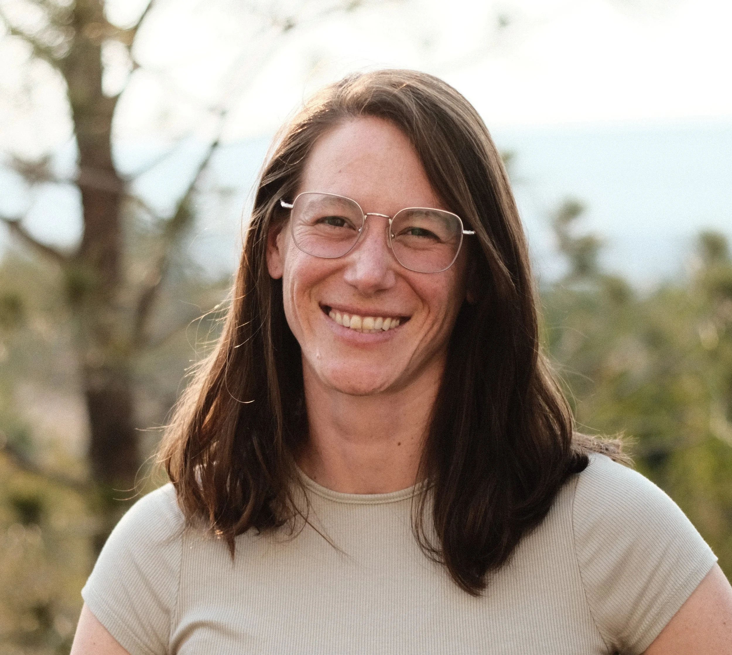 Close-up of a woman with long brown hair, wearing glasses and a light-colored shirt, smiling outdoors with blurred trees in the background.