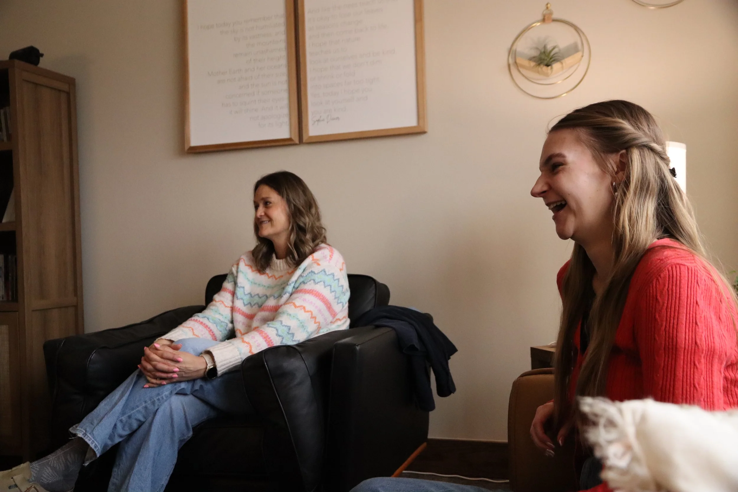 Two women with light skin and long hair sitting and laughing in a cozy room with framed pictures and a potted plant wall decor.