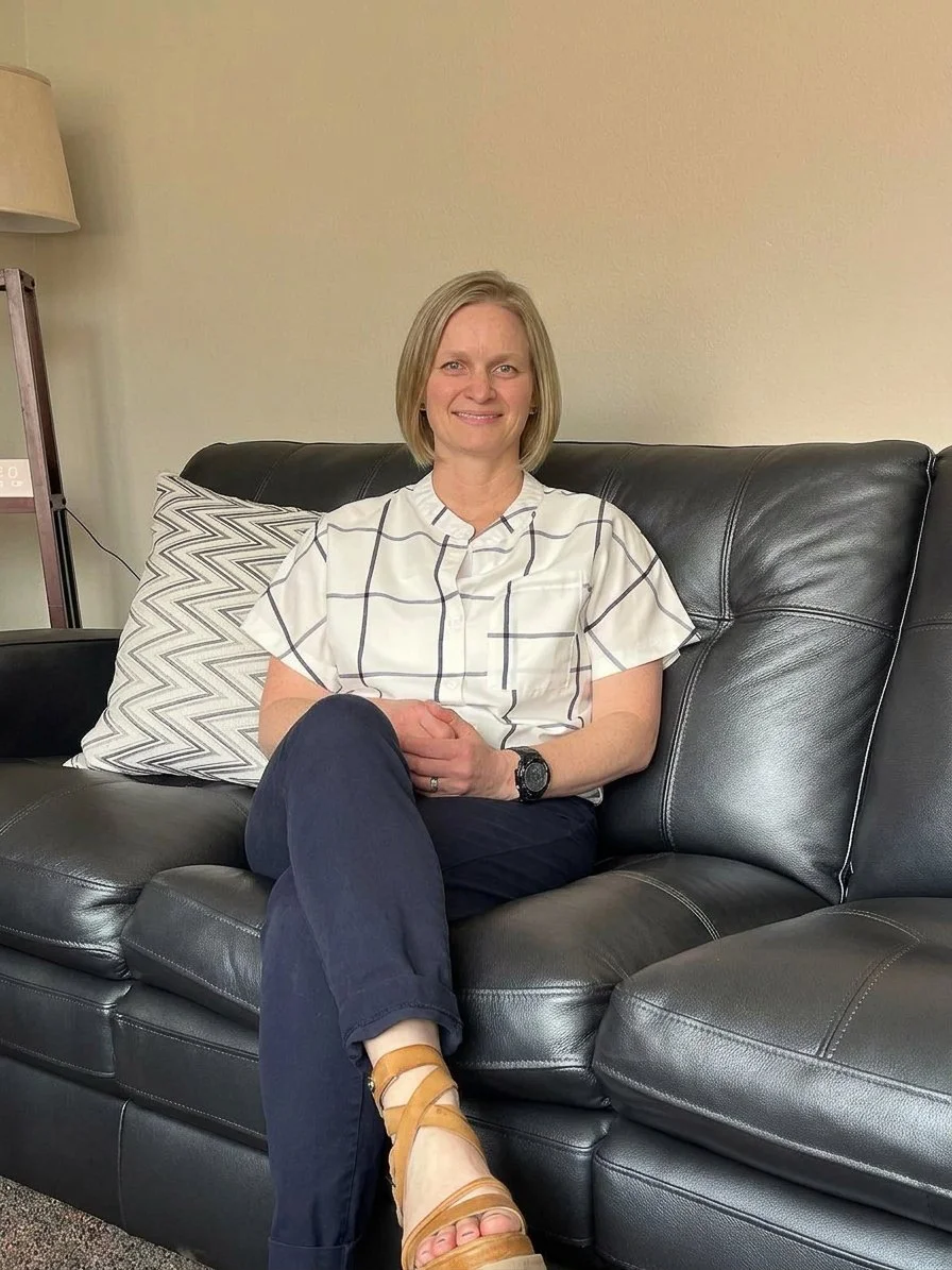 A woman with short blonde hair sitting on a black leather couch with a white and black checkered shirt and dark pants, smiling at the camera. There is a pillow with a geometric pattern next to her and a lamp and bookshelf in the background.