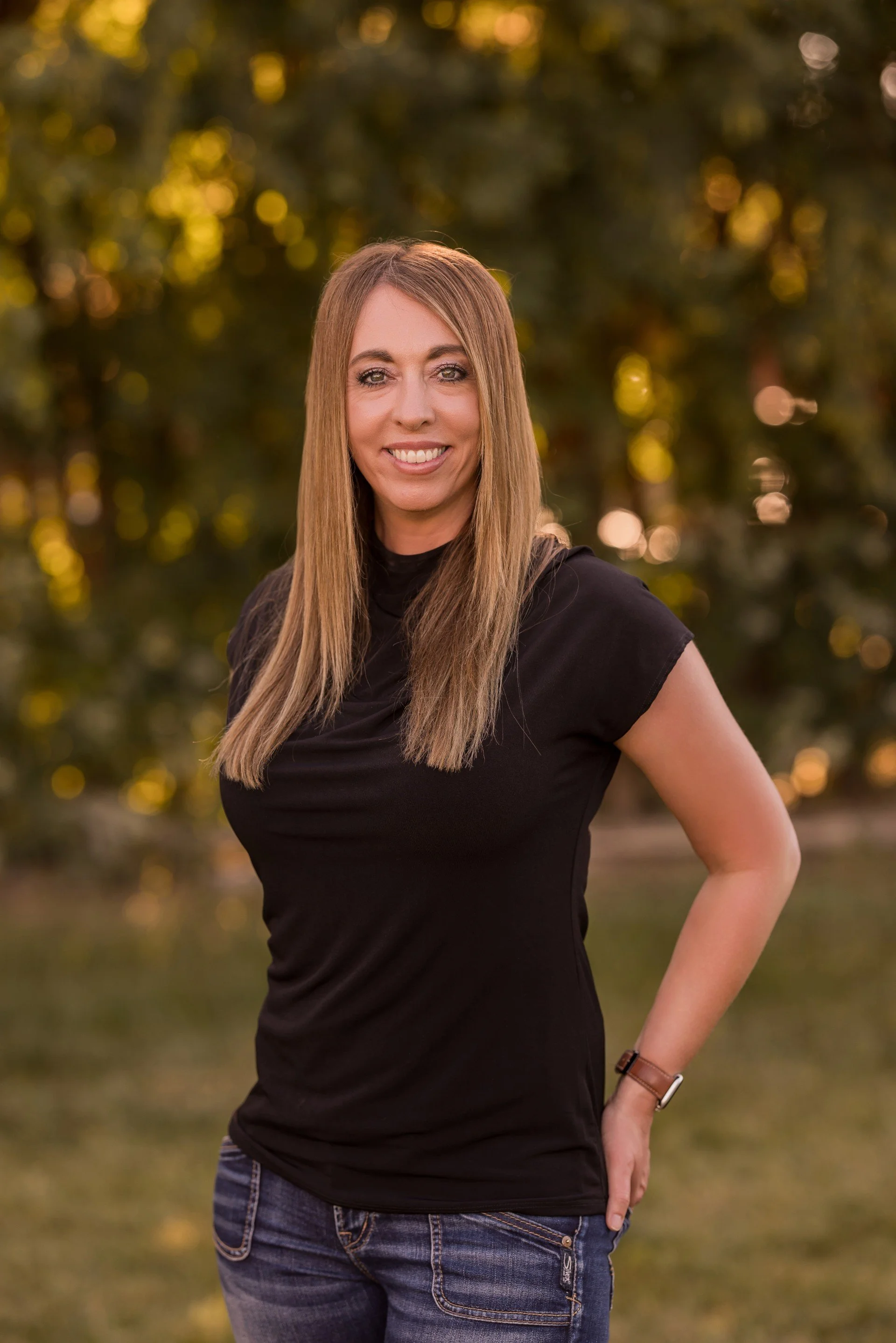 A woman with long blonde hair, wearing a black sleeveless top, jeans, and a watch on her left wrist, standing outdoors with trees and sunlight in the background.