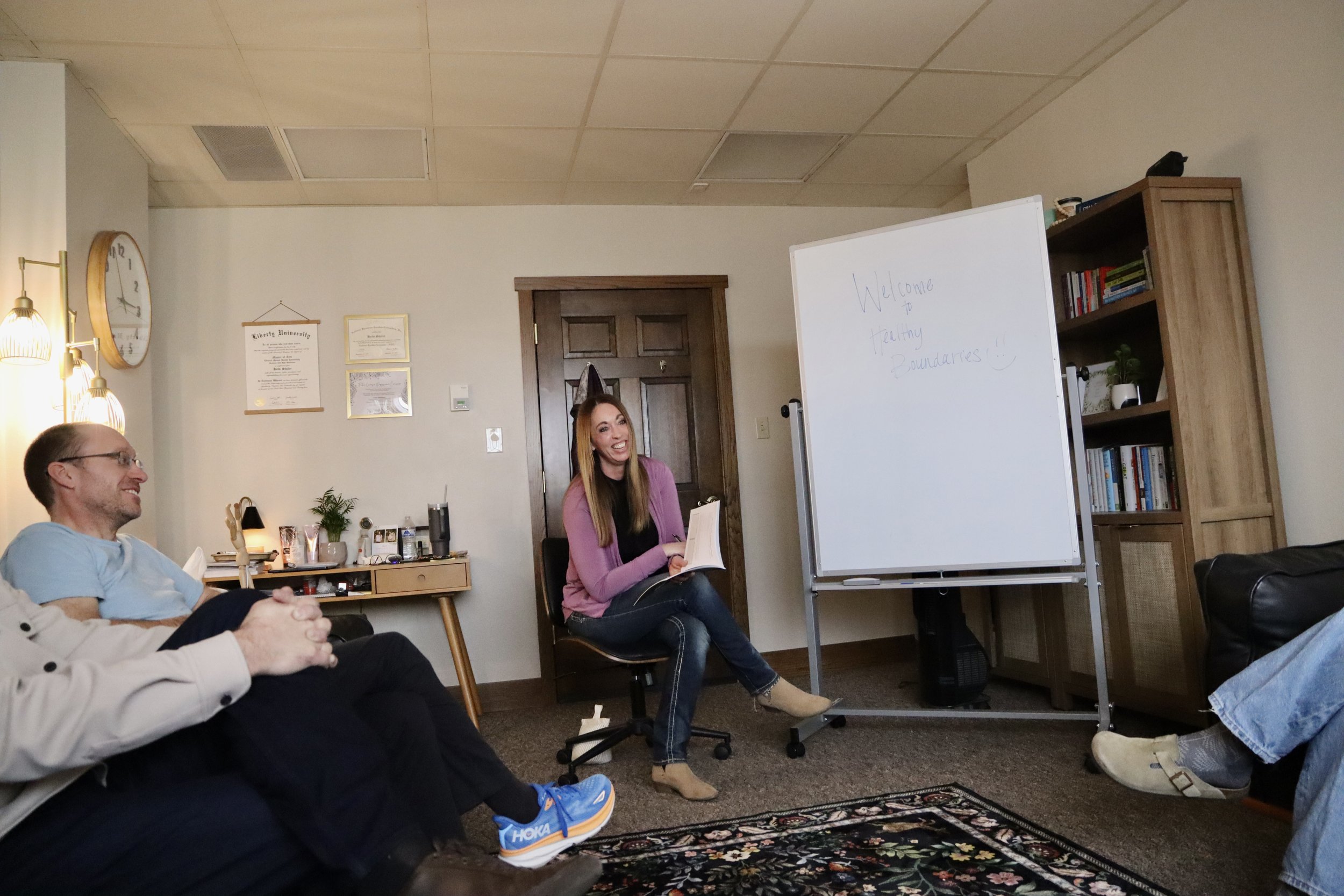 A female siting in a chair holding a book with a white board on the side.  Feet are visible from other individuals in the room.