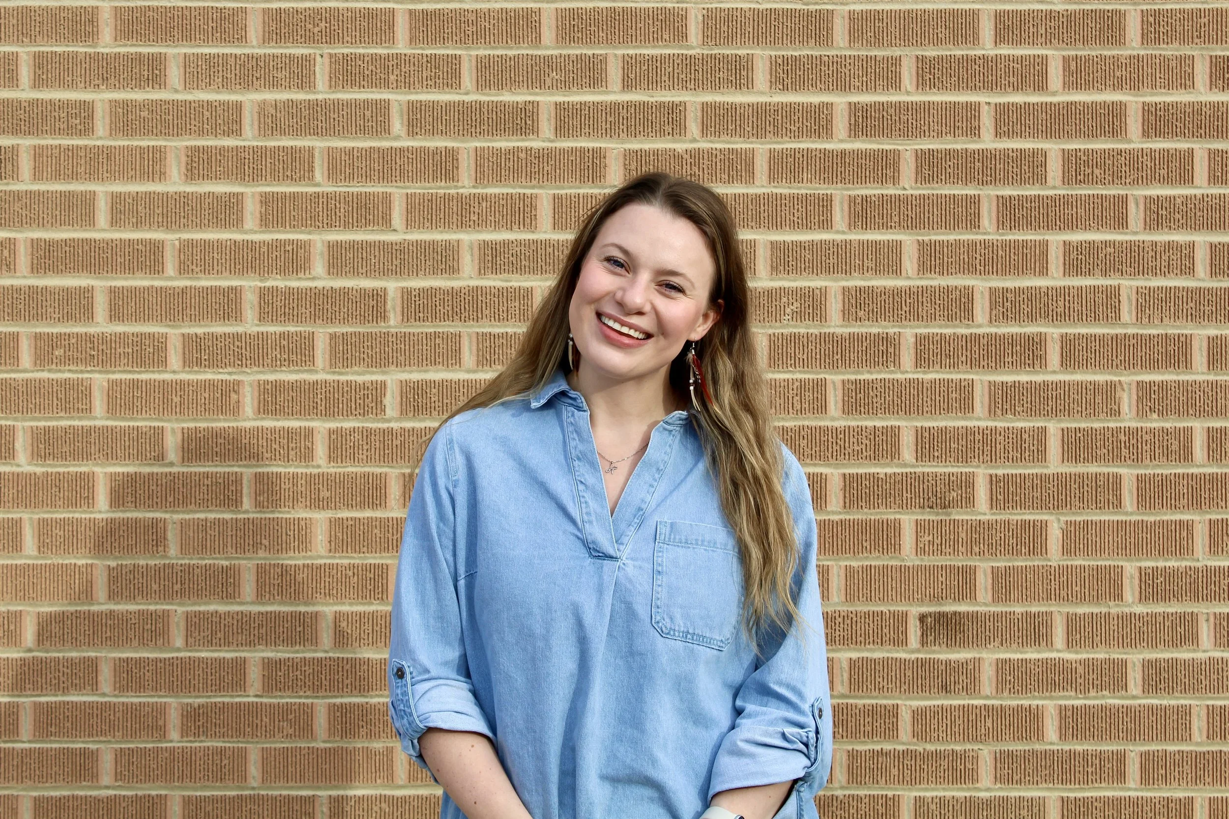 A woman in a blue denim shirt stands in front of a brick wall, smiling at the camera.