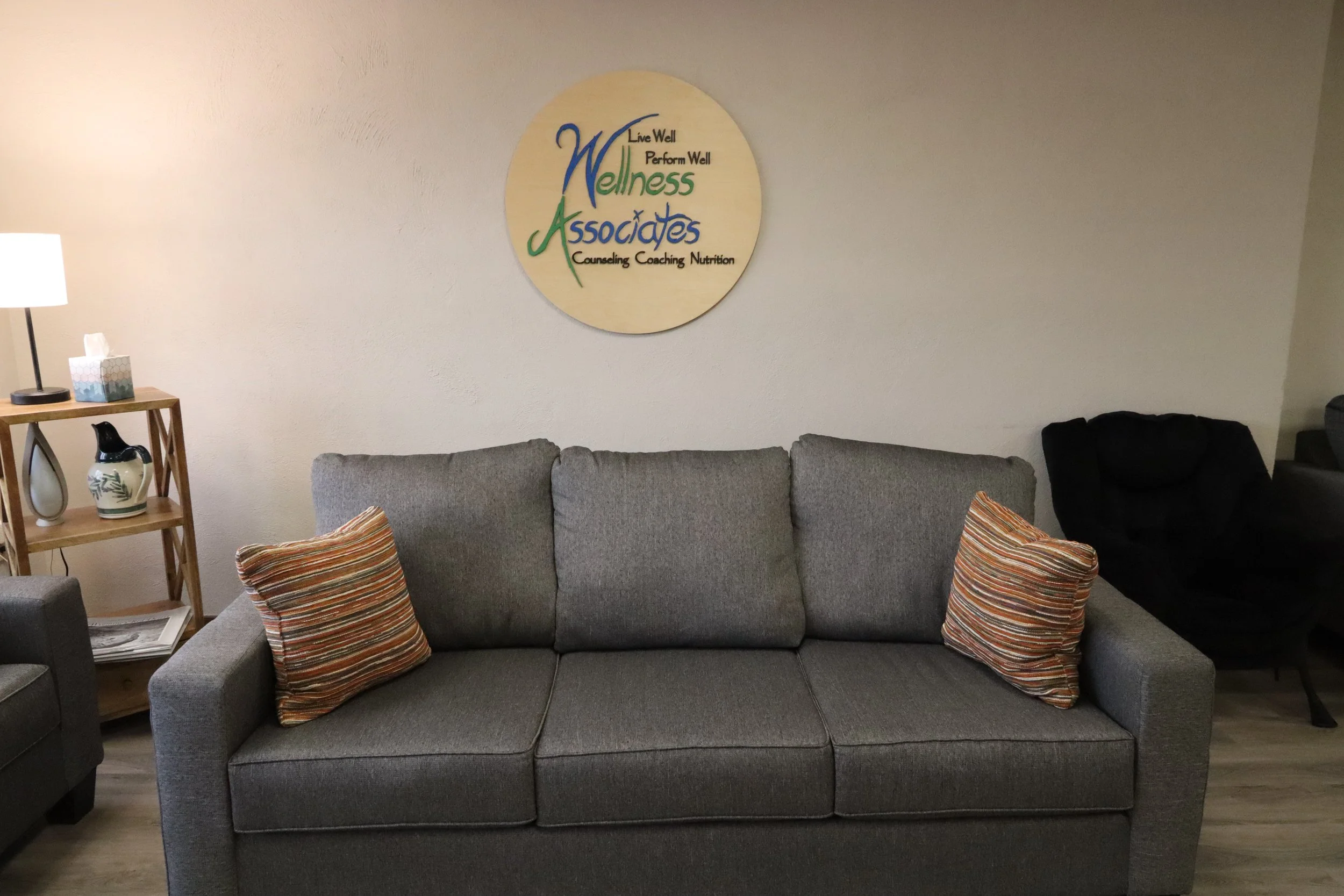 The living room wall with a circular sign for Wellness Associates, with the words 'Live Well, Perform Well, Wellness, Associates, Counseling, Coaching, Nutrition'. A gray sofa with three striped pillows is in front of the wall, and a side table with a lamp, tissue box, and decorative vase is on the left. A black armchair is on the right. The floor is wood.