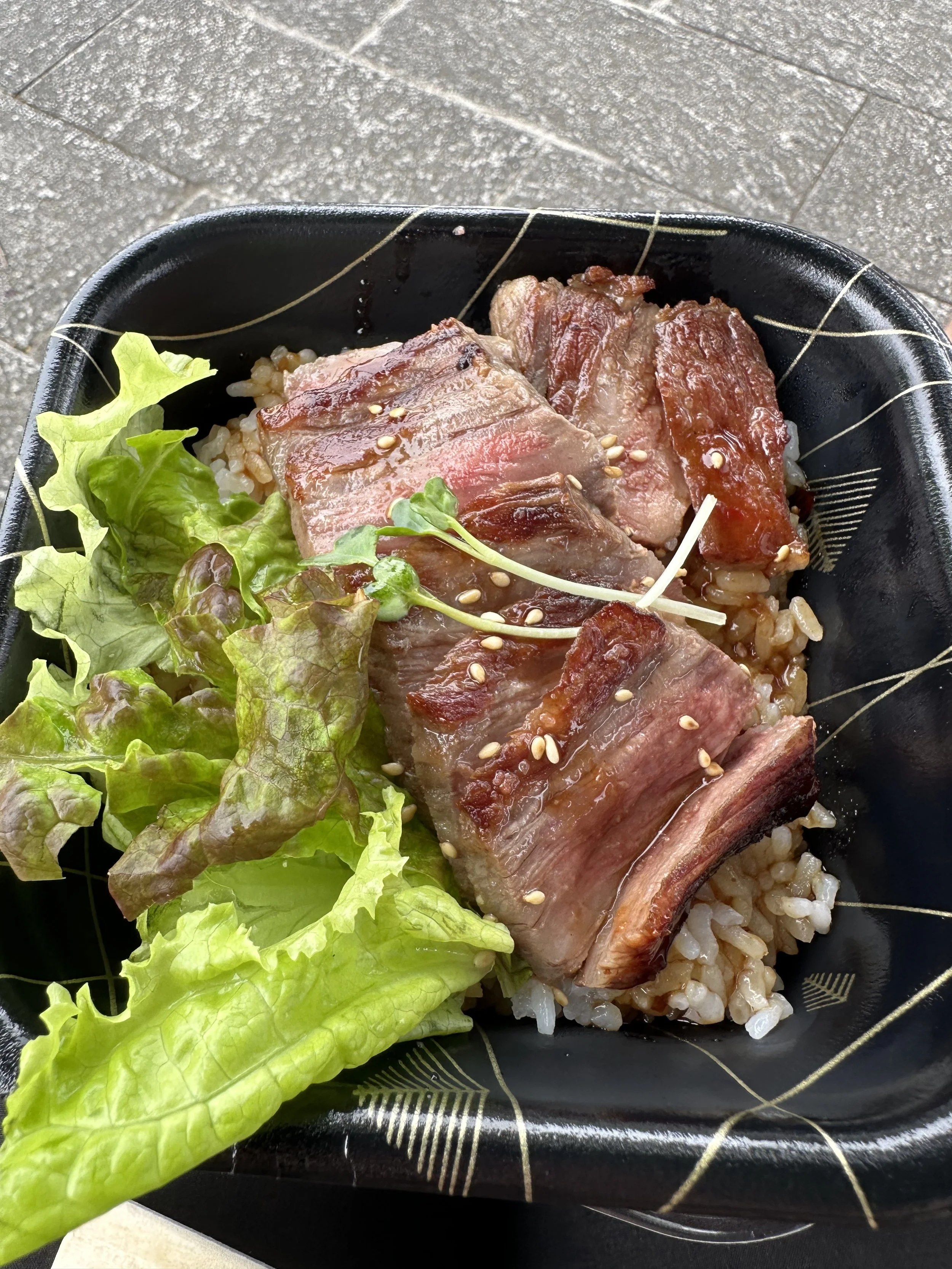 A black container with a bed of rice topped with slices of cooked beef, garnished with sesame seeds, and a side of leafy lettuce.