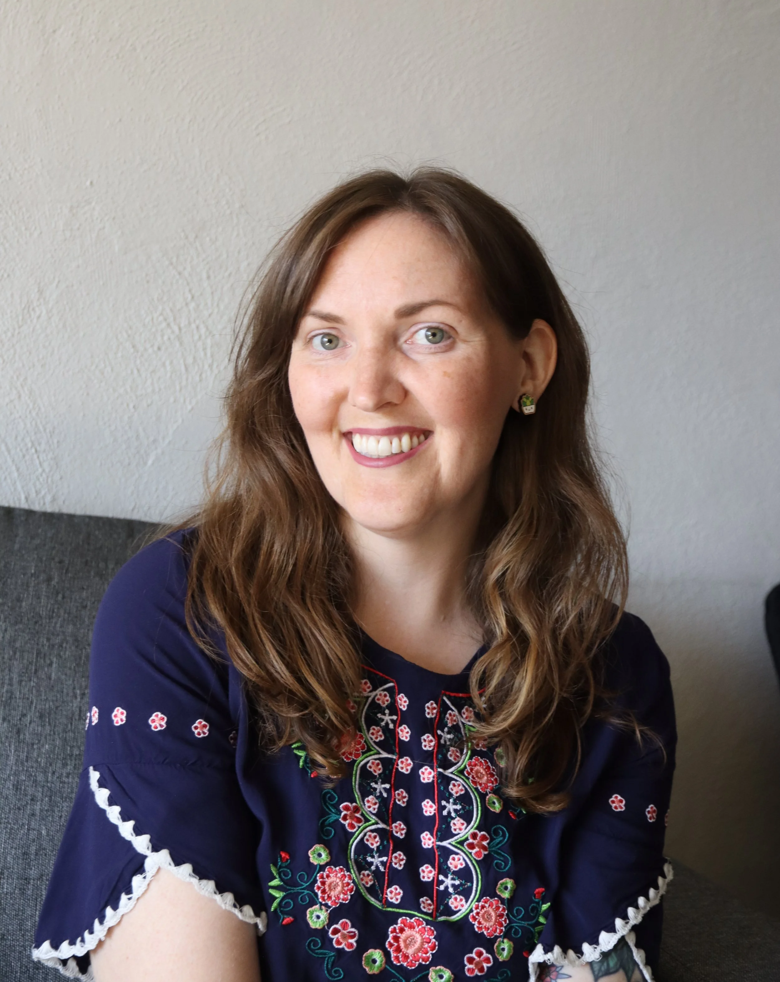 Close-up portrait of a woman with light skin, shoulder-length brown hair, and green eyes, smiling in front of a white textured wall. She is wearing embroidered earrings and a navy blue top with colorful floral embroidery.