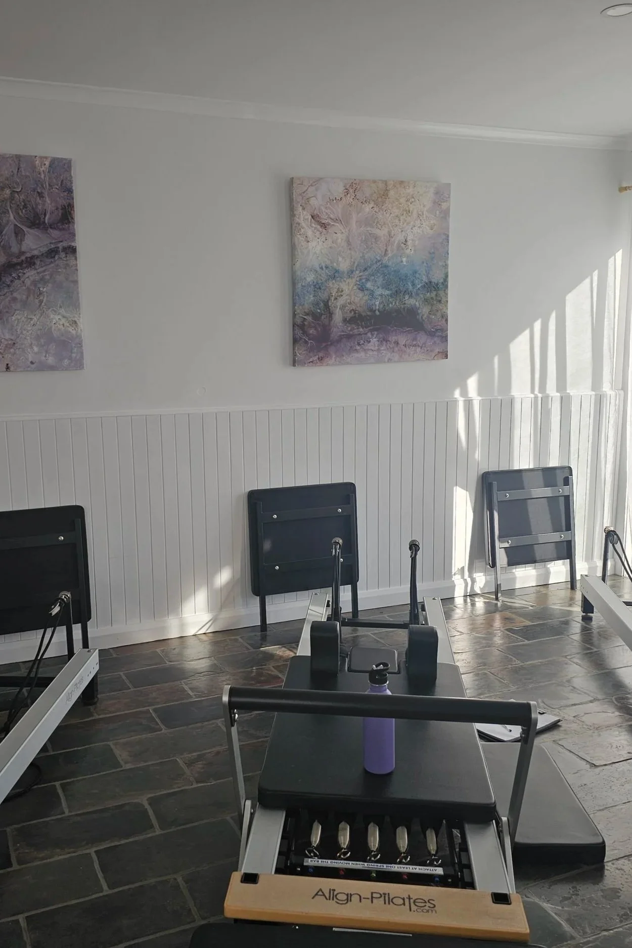 A Pilates class room with chairs against a white wall, two abstract paintings, and Pilates reformer equipment in the foreground with an Align-Pilates box, water bottle, and fitness accessories on top.