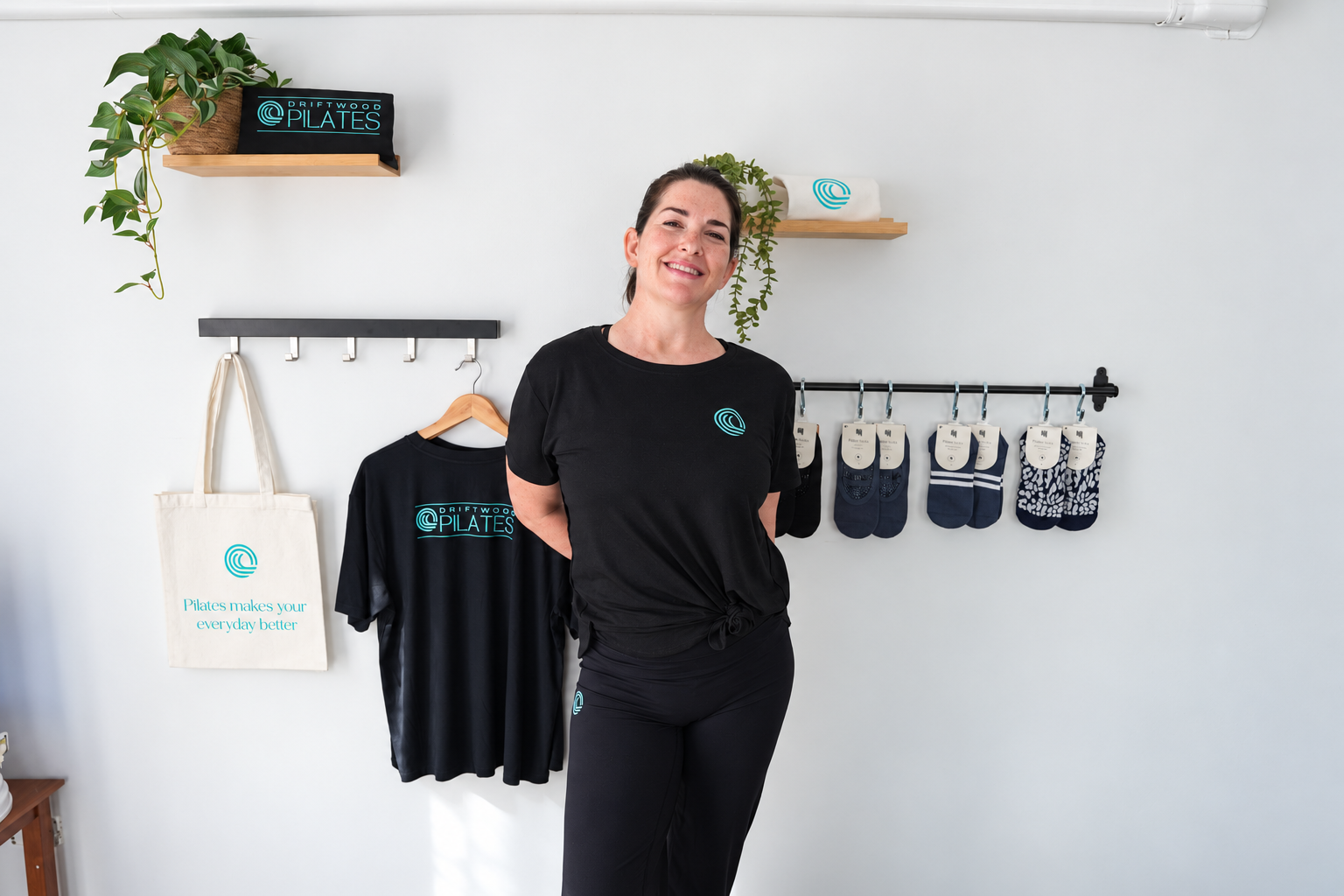 A woman in black workout clothing is using Pilates equipment with pulleys and straps. There are large exercise balls on shelves behind her, and windows with green trees outside.