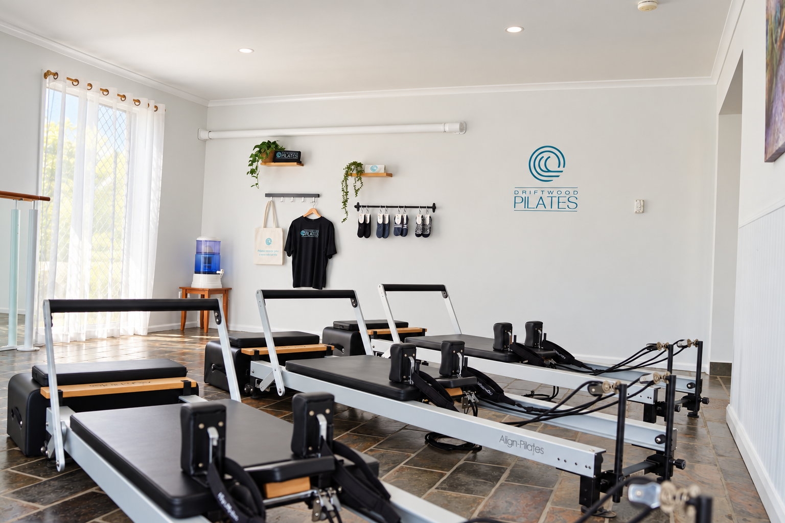 A Pilates class room with chairs against a white wall, two abstract paintings, and Pilates reformer equipment in the foreground with an Align-Pilates box, water bottle, and fitness accessories on top.