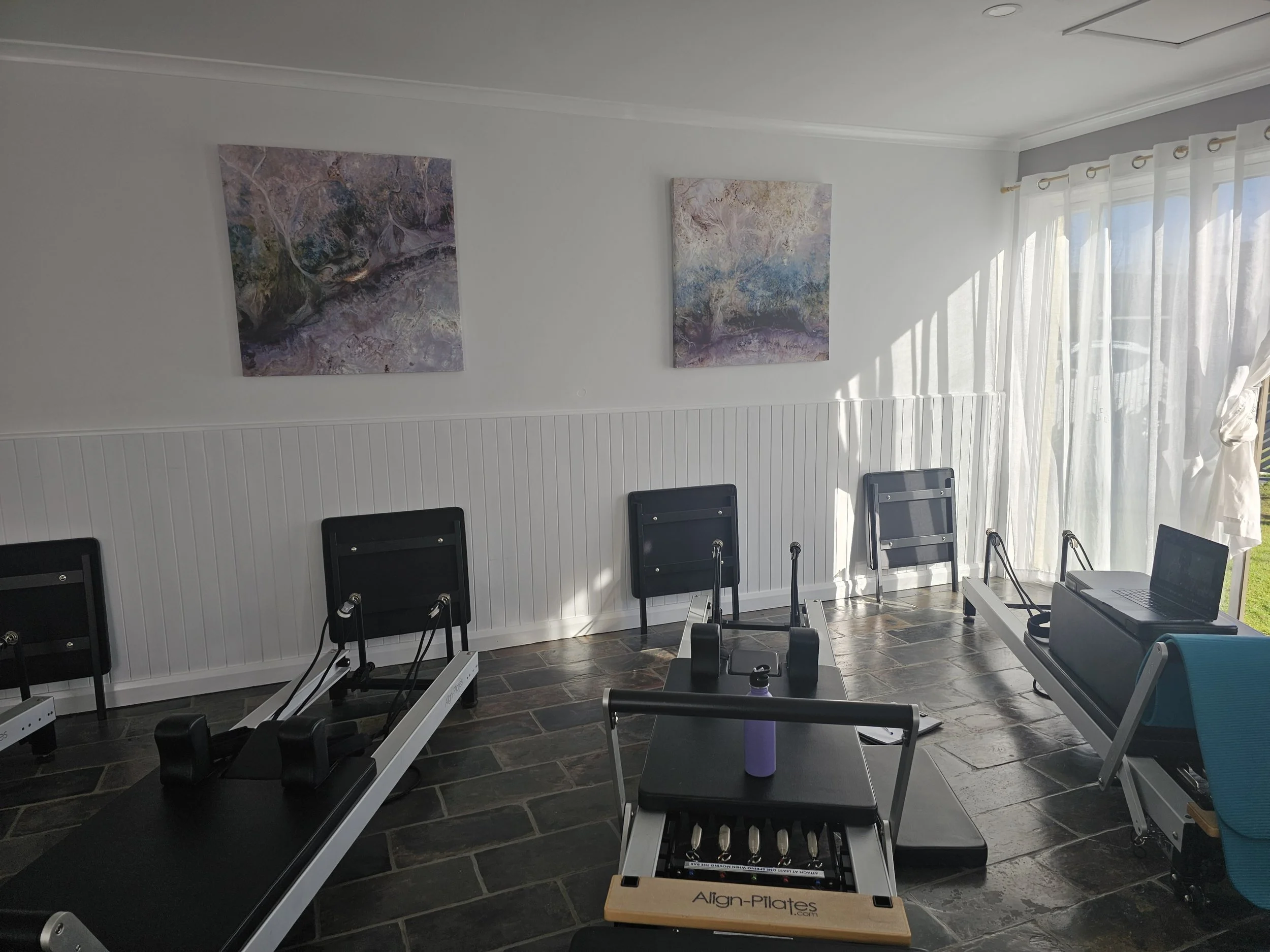 A room with white walls and sheer curtains letting in natural light. On the wall, there are two abstract paintings with purple, blue, and neutral tones. Several black chairs are lined up along the wall, and fitness or Pilates equipment is visible, including reformers and a wooden Pilates pillar with an