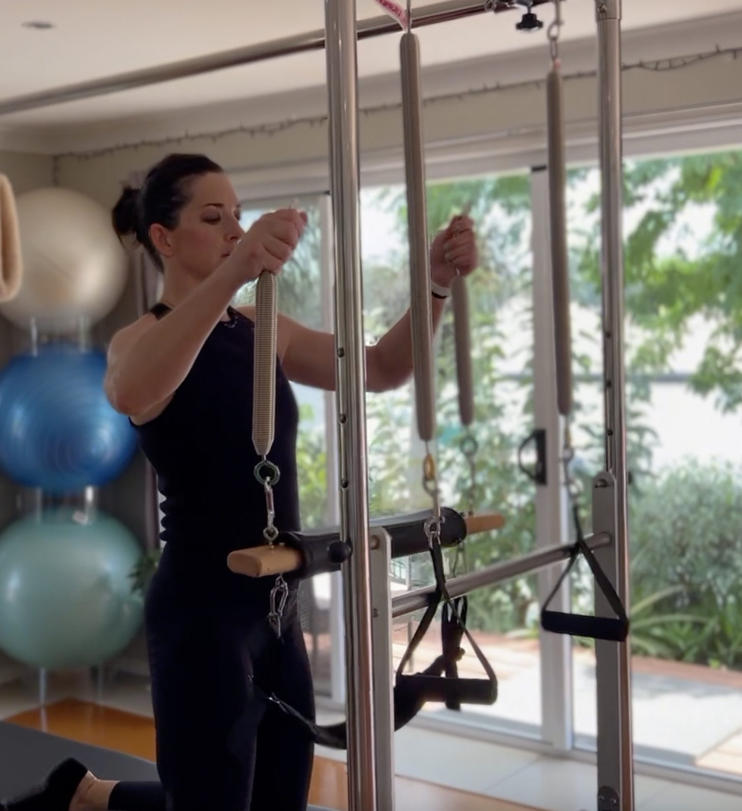 A woman in black workout clothing is using Pilates equipment with pulleys and straps. There are large exercise balls on shelves behind her, and windows with green trees outside.