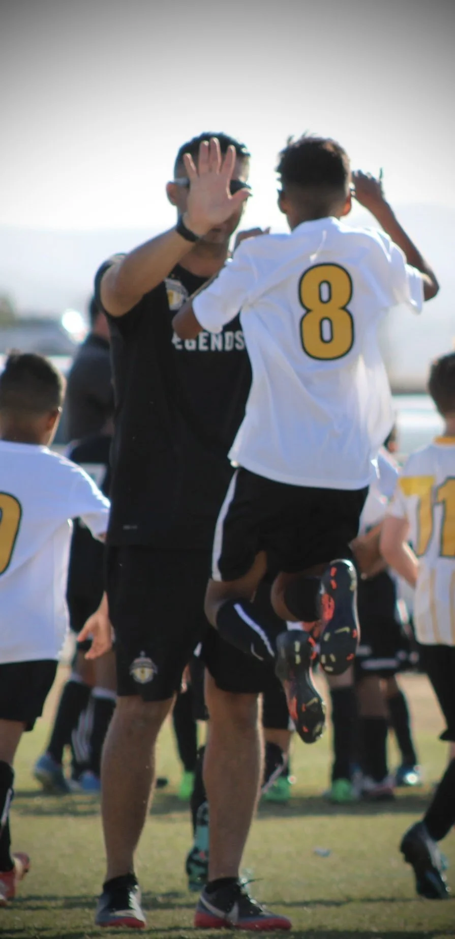 A coach lifting a young boy wearing a sports jersey with the number 8 during a youth soccer event outdoors on a sunny day.