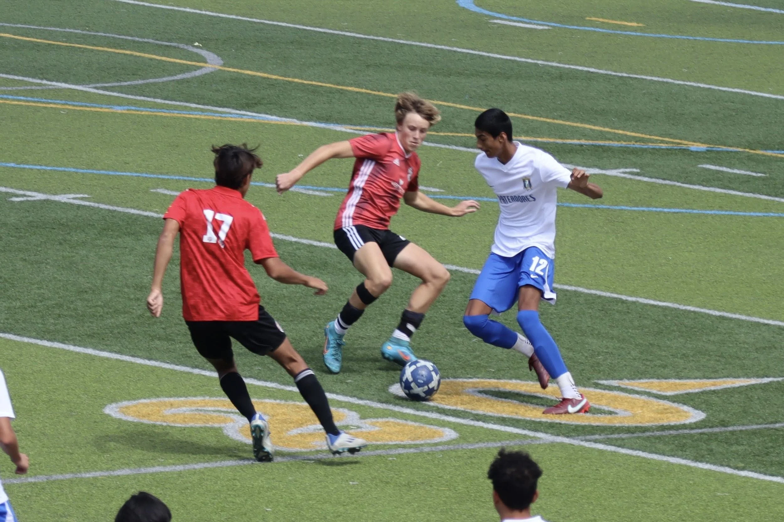 Two soccer players in red uniforms and one in white uniform compete for the ball on a green field with colorful lines.