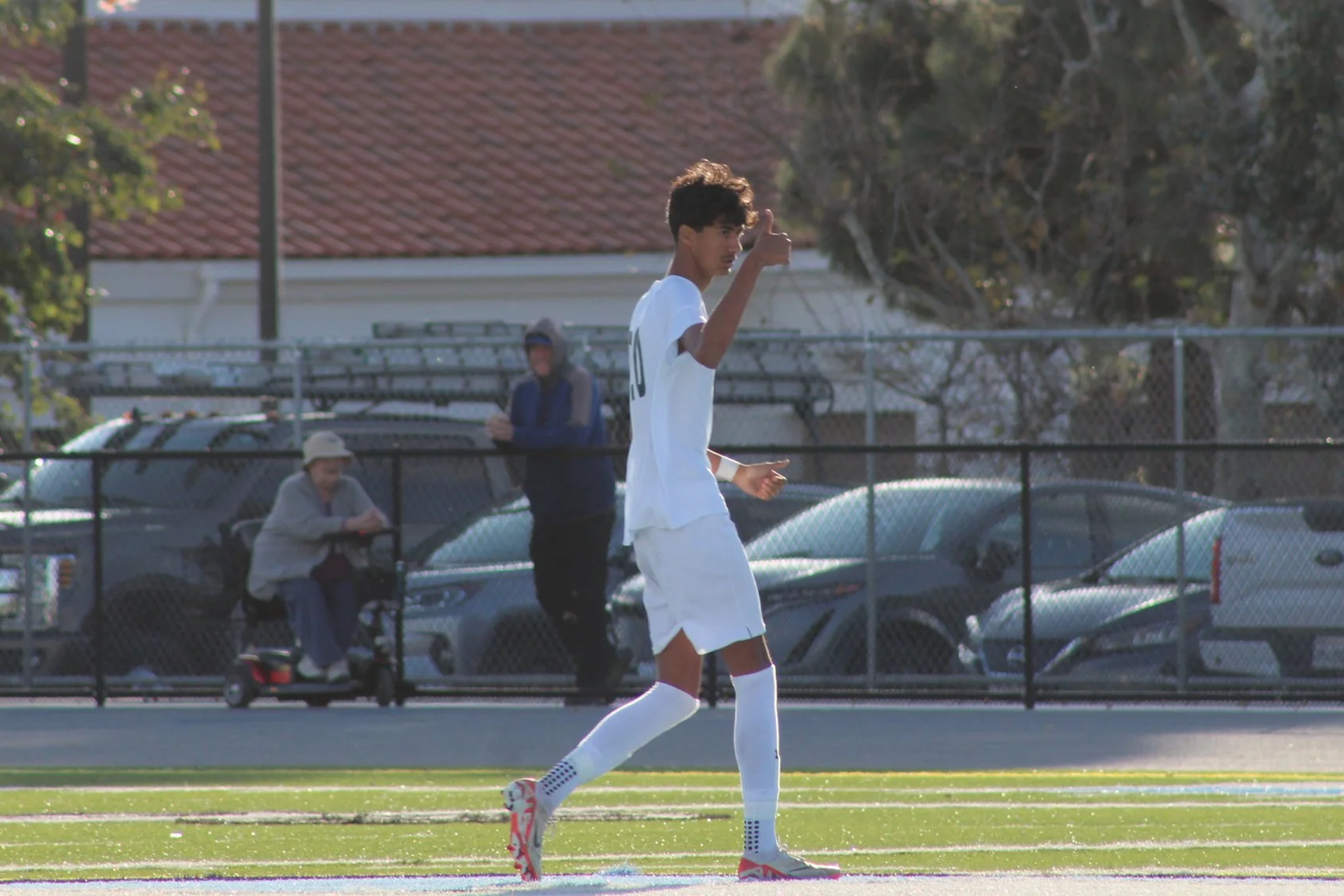 A young male soccer player in white uniform giving a thumbs up while jogging on the field during a game, with spectators and parked cars in the background.