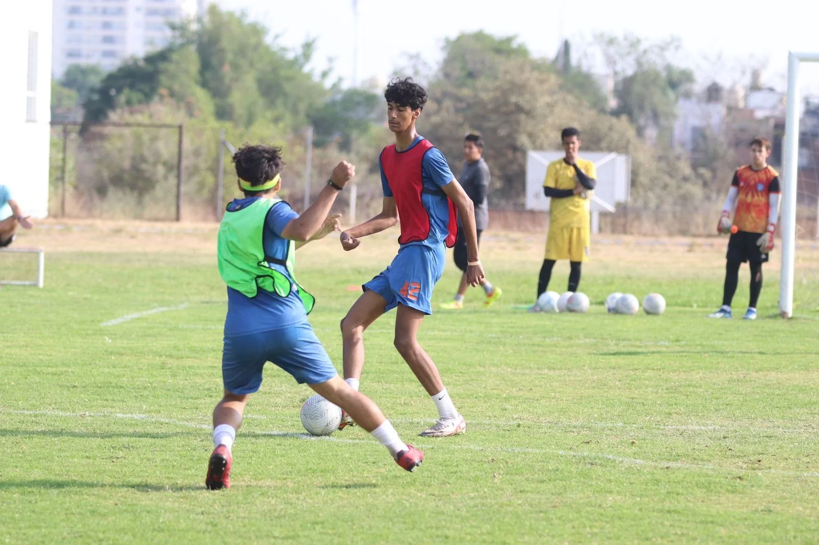 Two boys play soccer on a grassy field, one in a green vest and the other in red and blue sportswear, with a soccer ball between them. Several other players and coaches are visible in the background.