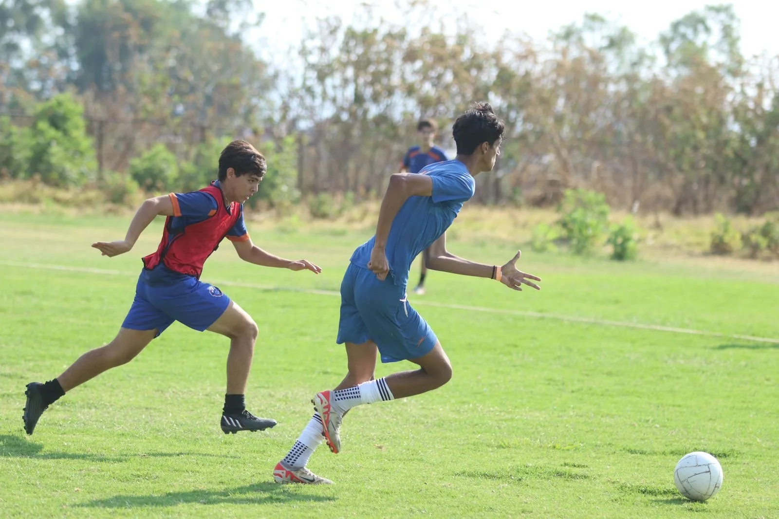 Two young soccer players running on a green field chasing a soccer ball, with trees in the background.