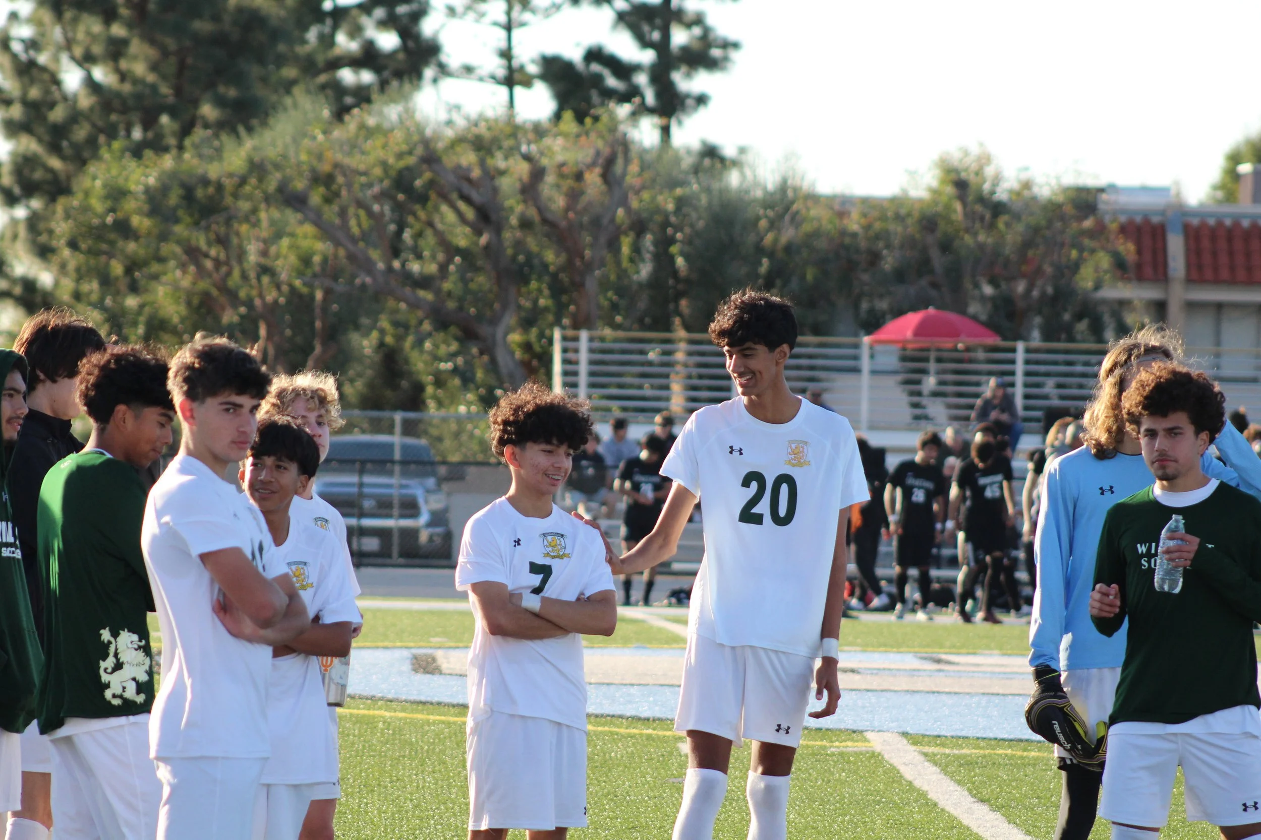 Group of young male soccer players standing on a field, wearing uniforms, during a match or practice, with trees and spectators in the background.