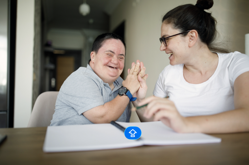 Two women sitting at a table, smiling and engaging in a joyful moment, with a notebook and pen on the table in a home setting.
