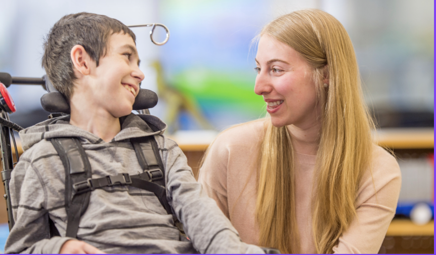 A young boy in a wheelchair sharing a joyful moment with a woman, both smiling at each other in an indoor setting.