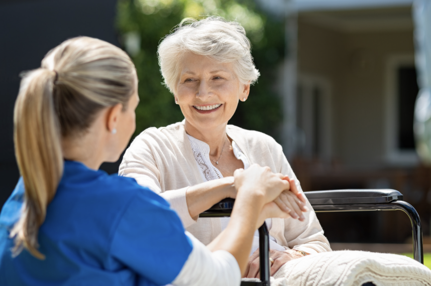 An elderly woman smiling and holding hands with a young caregiver outside on a sunny day, with a house in the background.