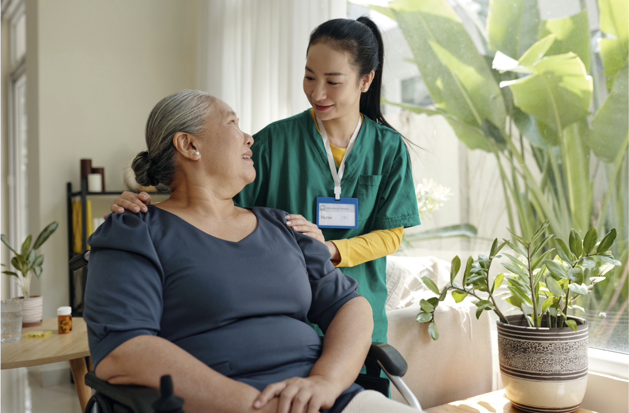A young healthcare worker in green scrubs and a name badge is gently holding the shoulders of an elderly woman with gray hair. They are indoors near a large window with sunlight and potted plants visible around.