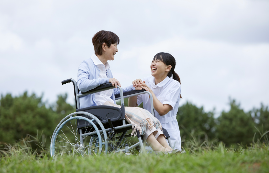 A young woman in a white uniform kneeling and holding hands with an elderly woman in a wheelchair outdoors on green grass, both smiling and laughing with a cloudy sky in the background.