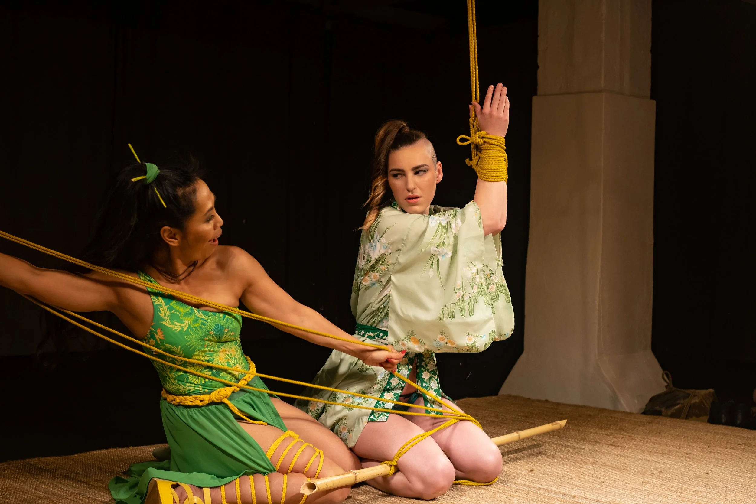Two women are kneeling on a mat with their hands and feet bound by yellow ropes. One woman in a green patterned dress has her arm raised with her palm open, while the other woman in a cream floral kimono-like outfit looks confused, with her hair partly shaved on one side.