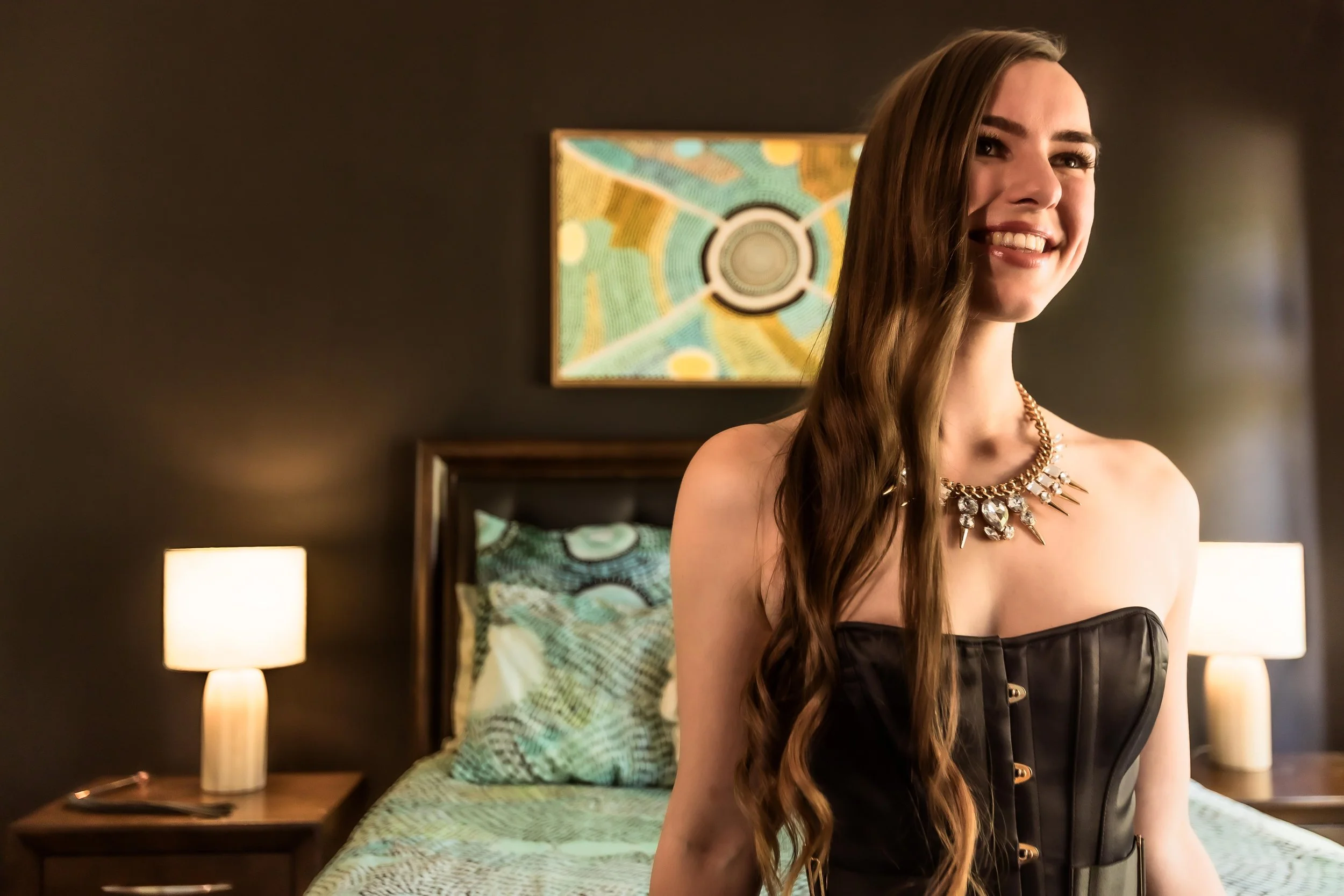 Young woman with long brown hair smiling, wearing a black strapless dress and a large statement necklace, sitting on a bed in a dimly lit hotel room with modern decor, including nightstands with lamps, a headboard, and colorful abstract artwork on the wall.