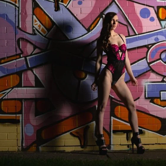 A woman with long dark hair wearing black and pink lingerie and high heels standing in front of colorful graffiti on a brick wall.