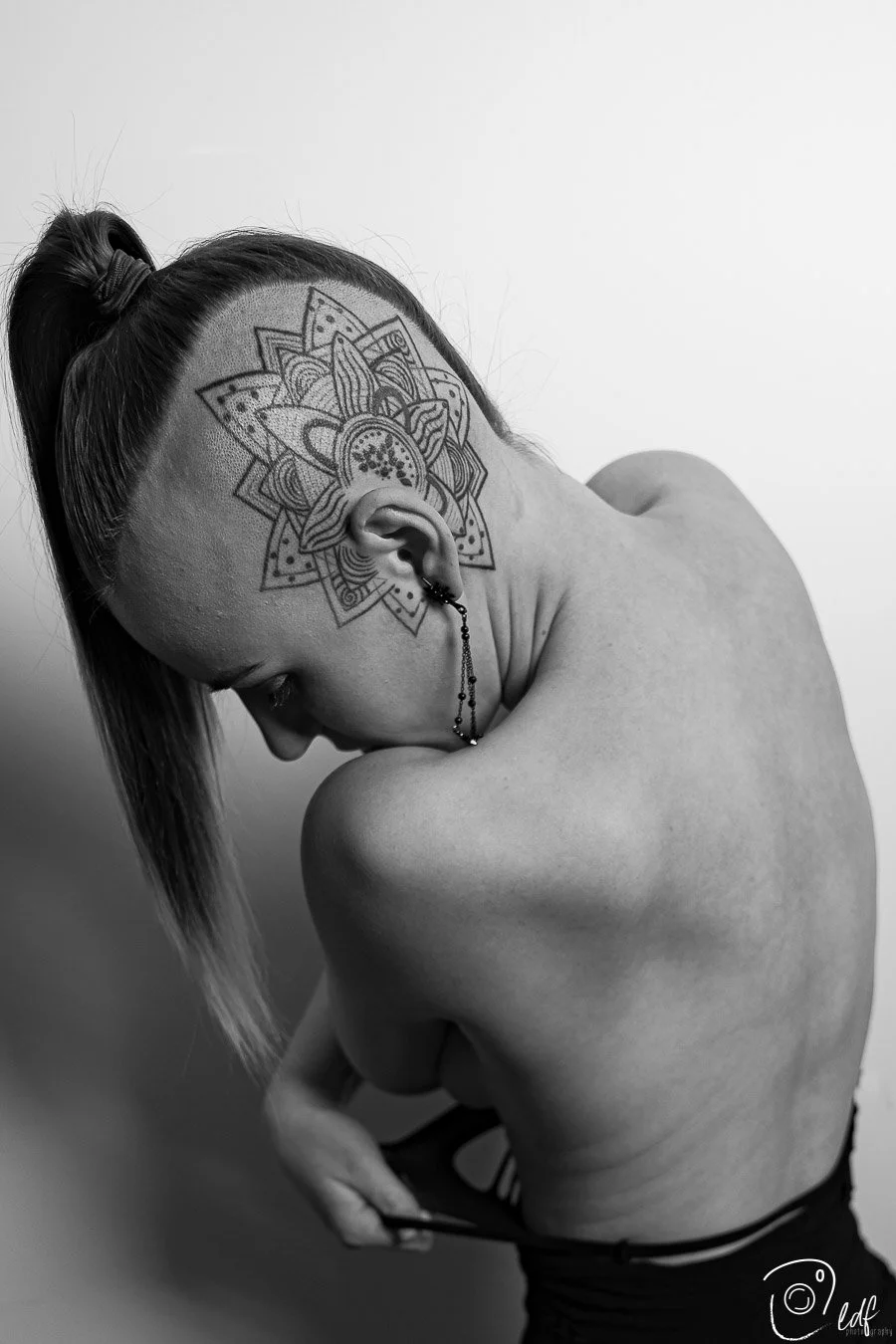 A black and white photo of a woman with a large, intricate tattoo on the side of her head and shaved hair on the side. She is looking down with her eyes closed and holding her clothing with one hand.