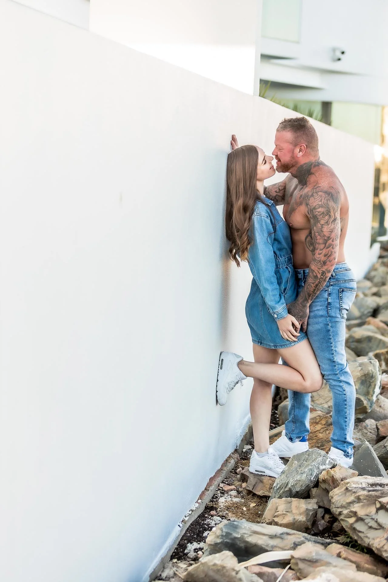 A couple stands on rocks by a white wall, sharing a close moment. The woman wears a denim jacket and white sneakers, with one leg raised and bent. The man is shirtless with tattoos, wearing jeans and white sneakers. They are leaning into each other with noses touching.