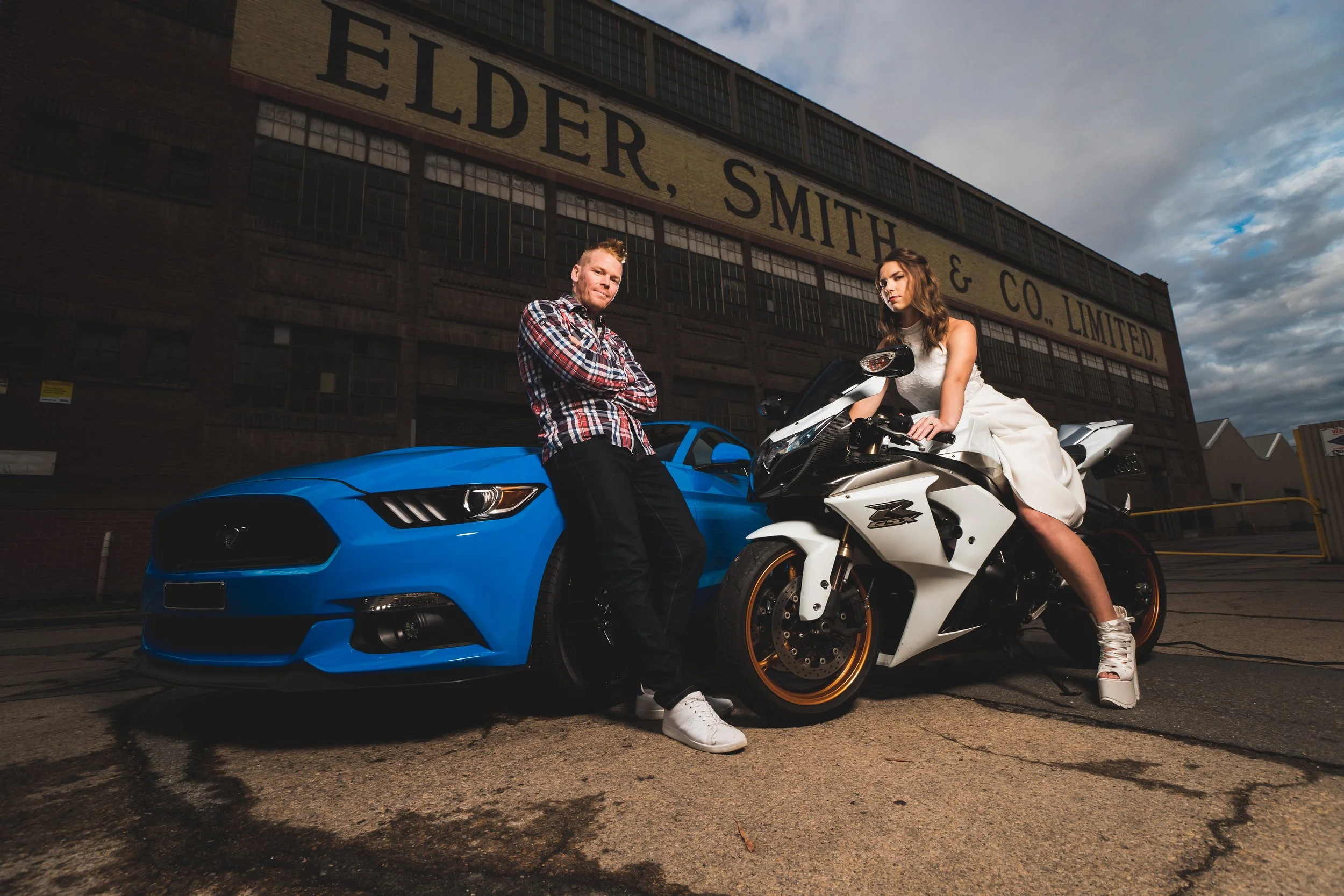 A man standing next to a blue sports car and a woman sitting on a white motorcycle in front of an industrial building with a sign saying Eldler, Smith & Co. Limited. The woman is wearing a white dress and white high-heeled shoes, and the man is wearing a plaid shirt and dark pants.