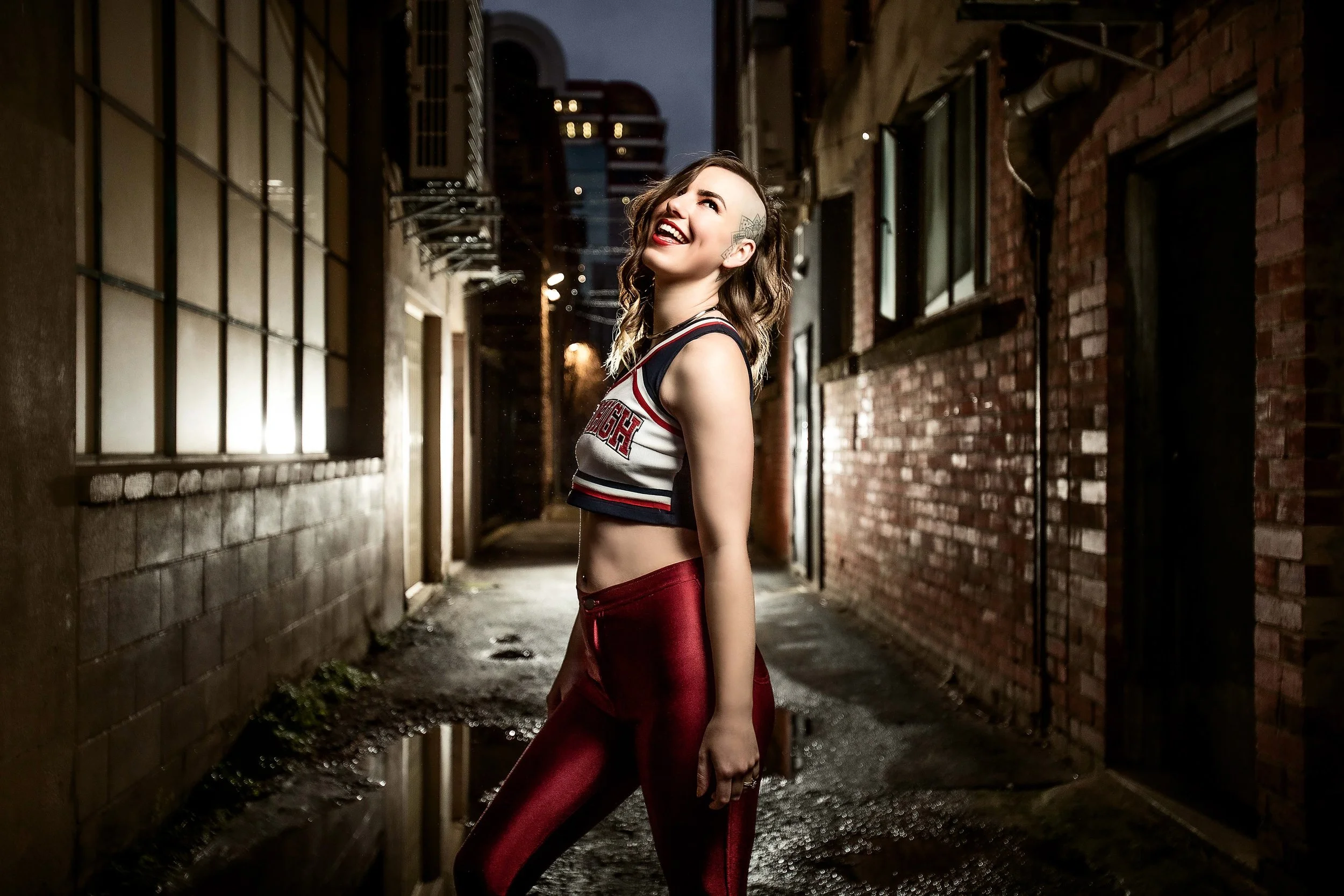 A smiling young woman with tattoos and wavy hair, wearing a cheerleading outfit and red shiny pants, standing in a dark alley at night with brick and window walls.